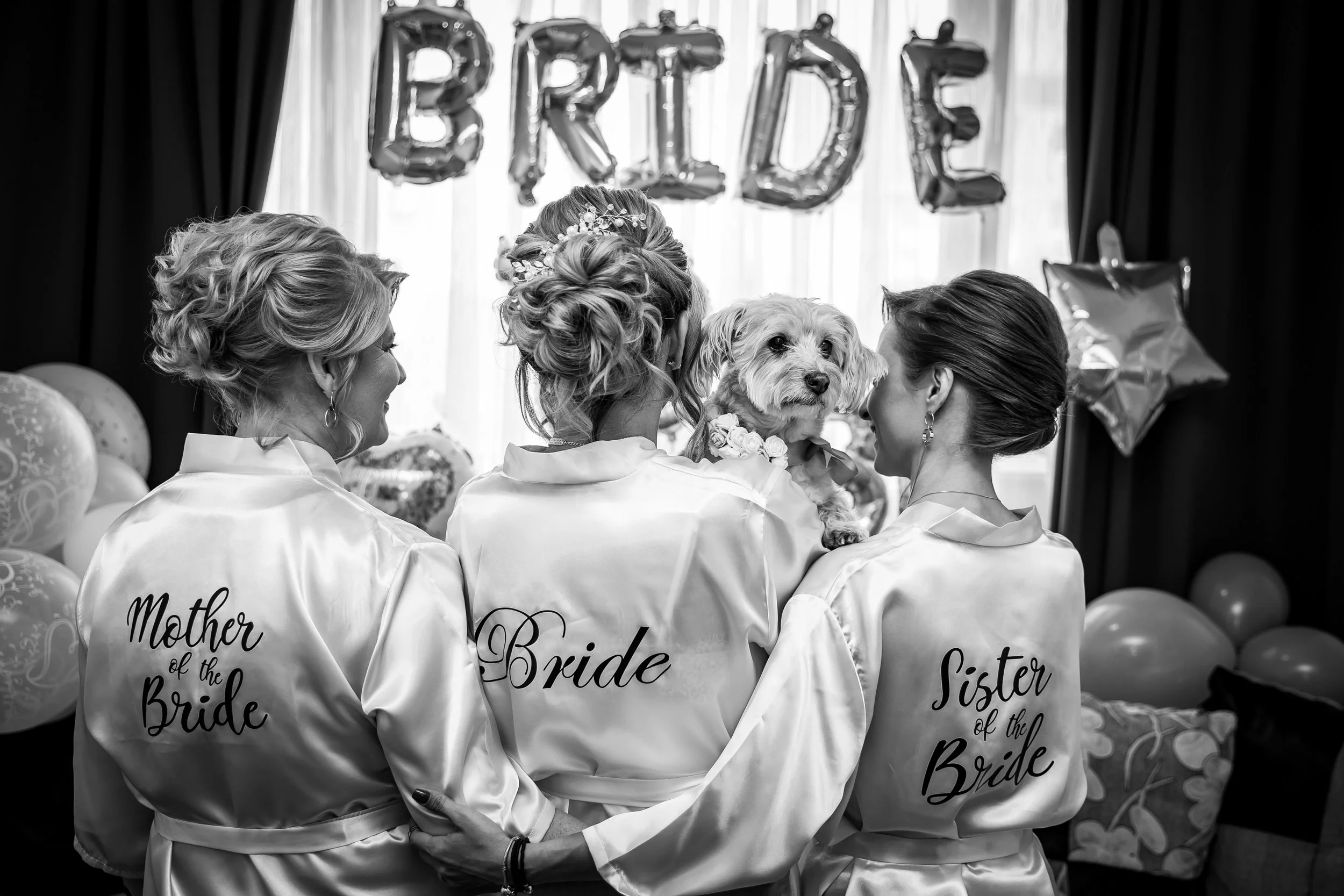Black-and-white photo of three women wearing matching satin robes with words like "Mother of the Bride," "Bridesmaid," and "Sister of the Bride" visible, standing close together and holding a small dog with a floral collar. There are balloons and lar