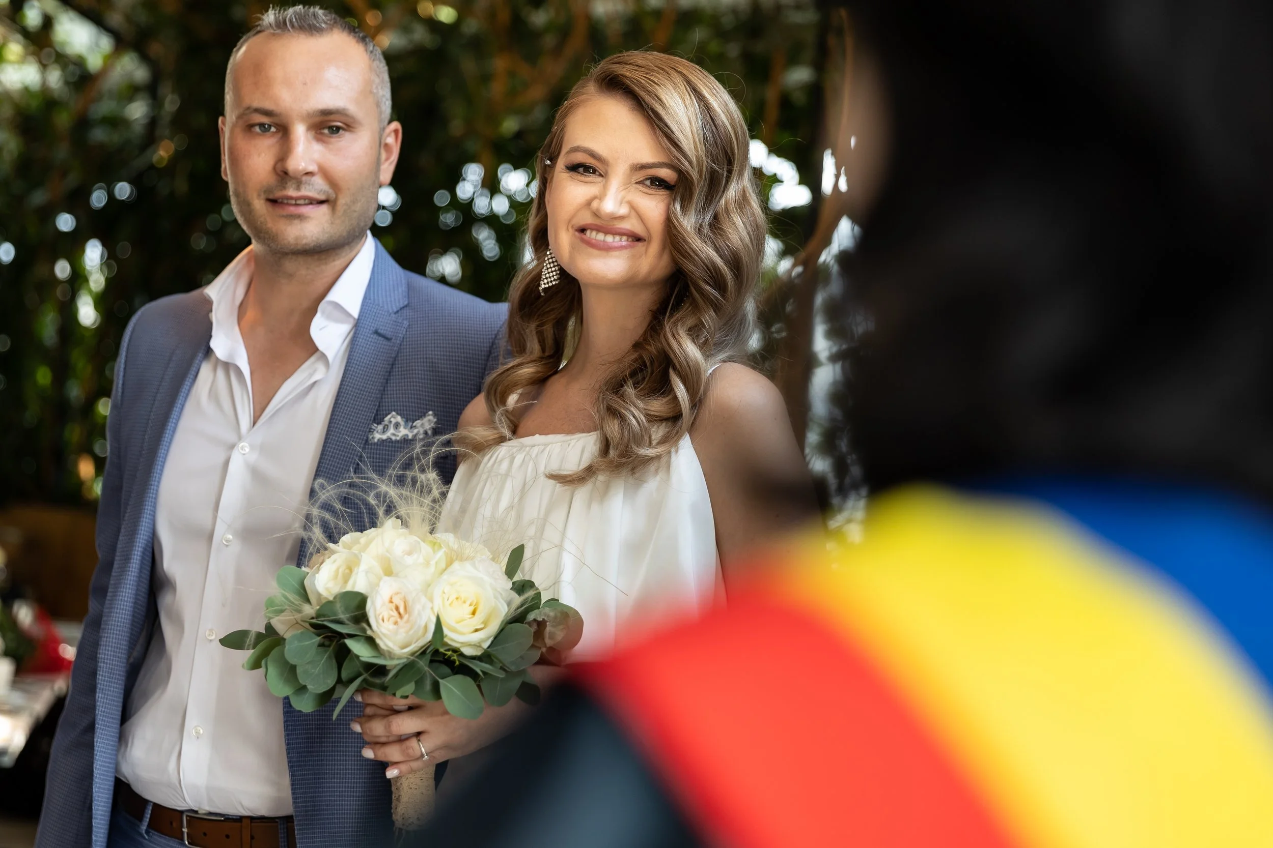 A bride holding a bouquet of white roses and greenery, standing next to a groom in a blue suit, both smiling during a wedding ceremony outdoors.