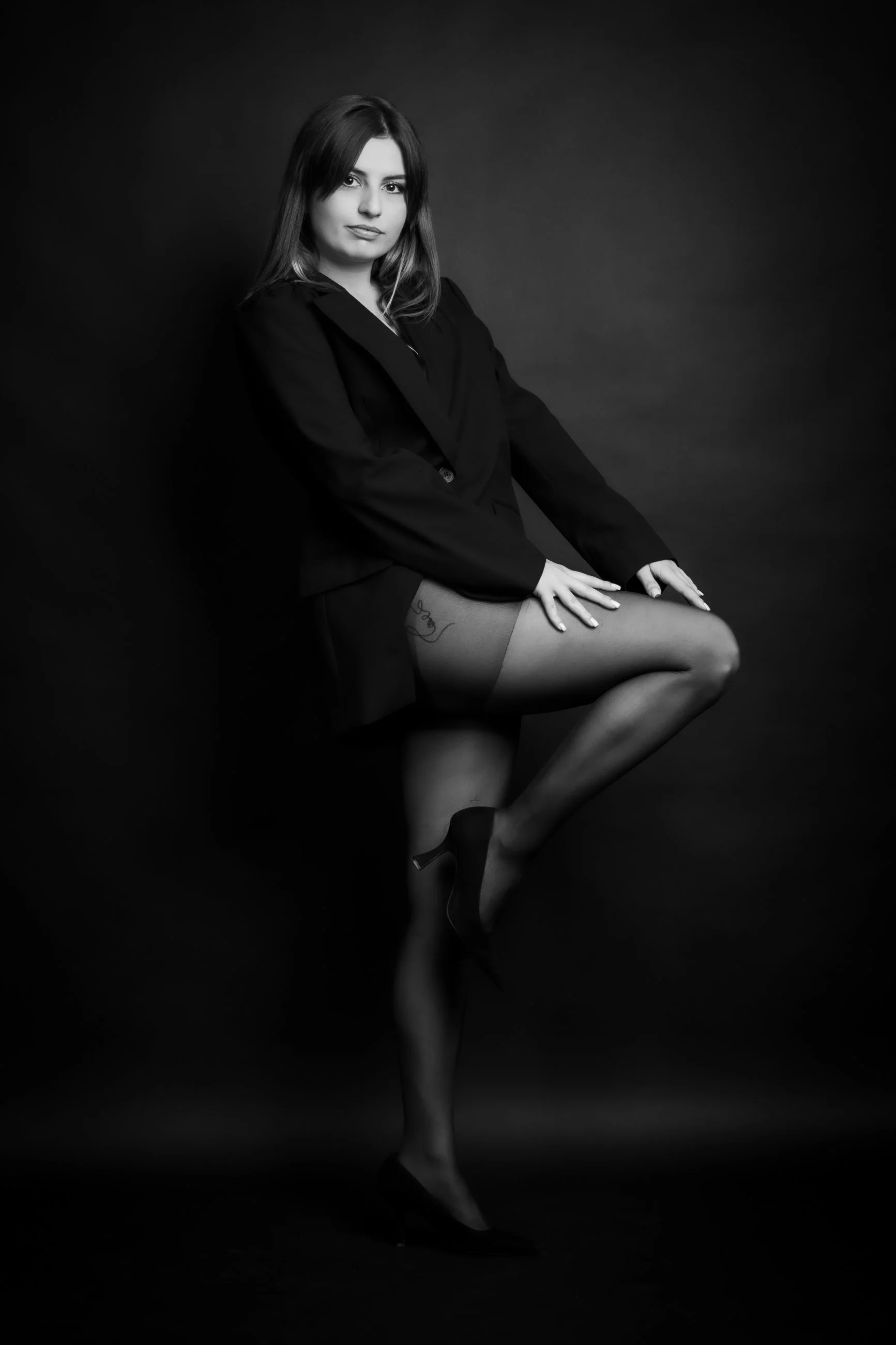Black and white photo of a woman sitting with her legs crossed, wearing a blazer, stockings, and heels, with a neutral expression against a dark background.
