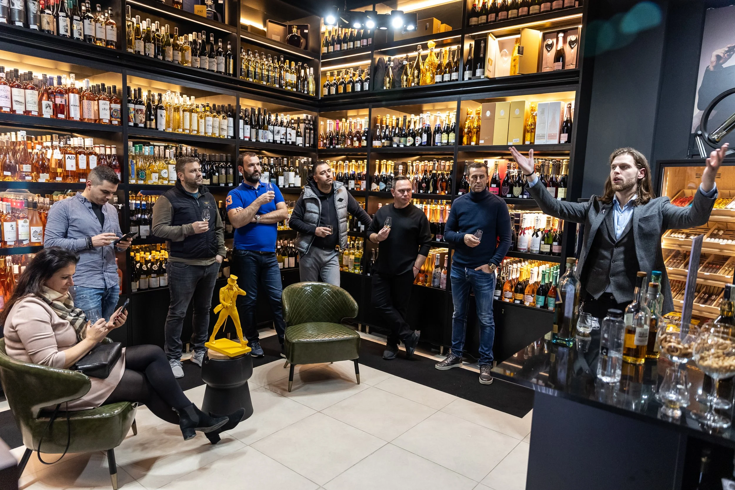 A wine tasting event in a wine store with shelves full of wine bottles. People are standing and sitting, some holding wine glasses. A man with long hair and a blazer is speaking and gesturing with his arms. Others are listening attentively, with one 