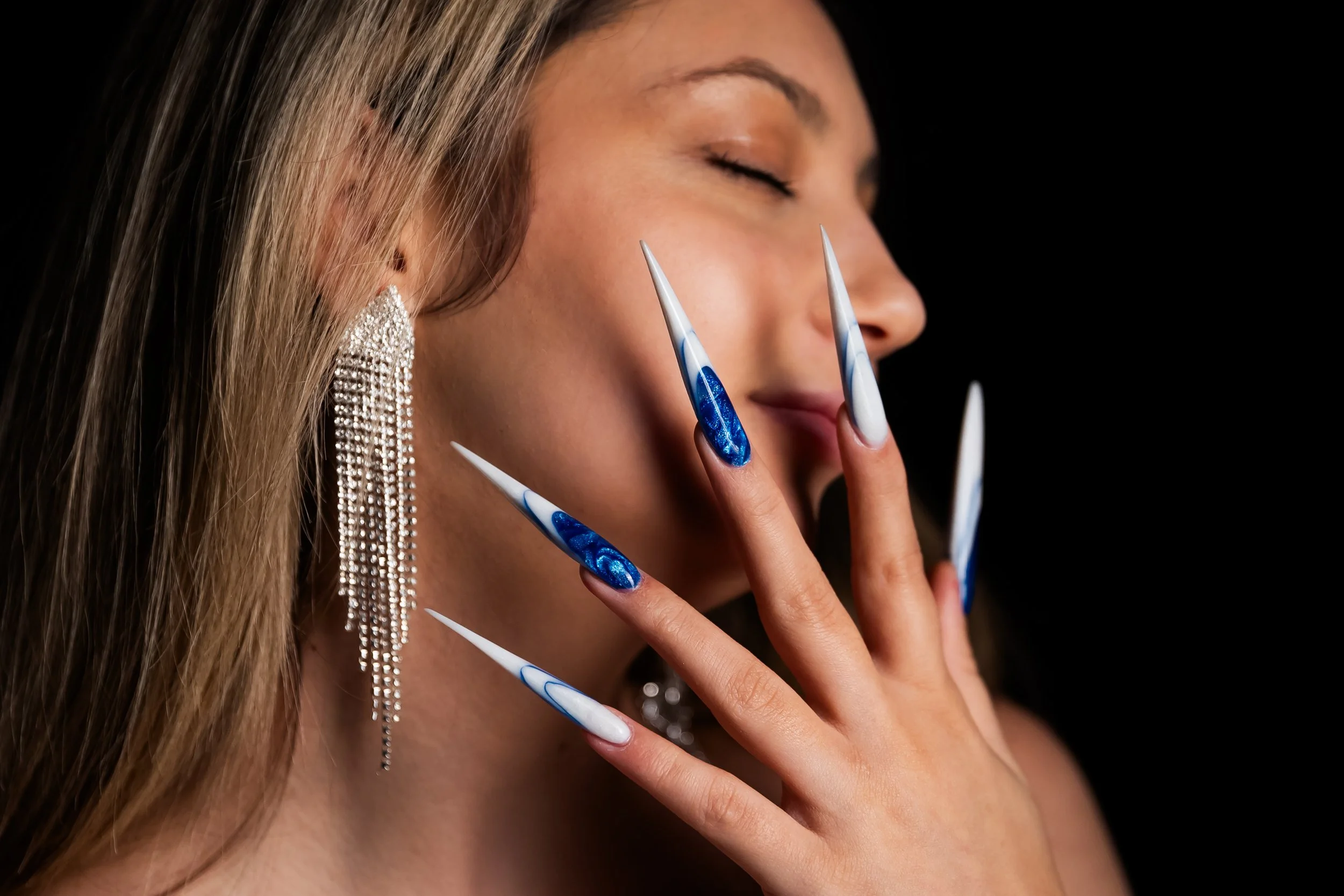 A woman with long, blonde hair and large, dangling diamond earrings has her eyes closed and is touching her face with her long, decorated nails, which are painted with blue and white designs.
