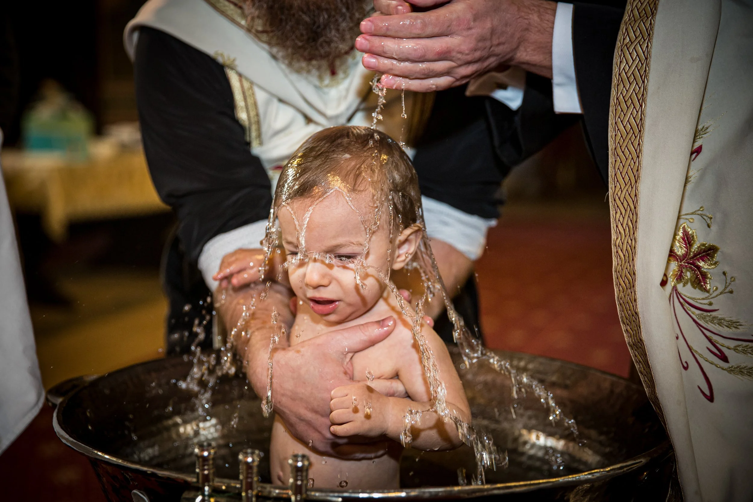 A young child with wet hair, standing in what appears to be a baptismal font, being baptized by a man in ceremonial robes by pouring water over their head. The child's facial expression shows focus or slight discomfort.