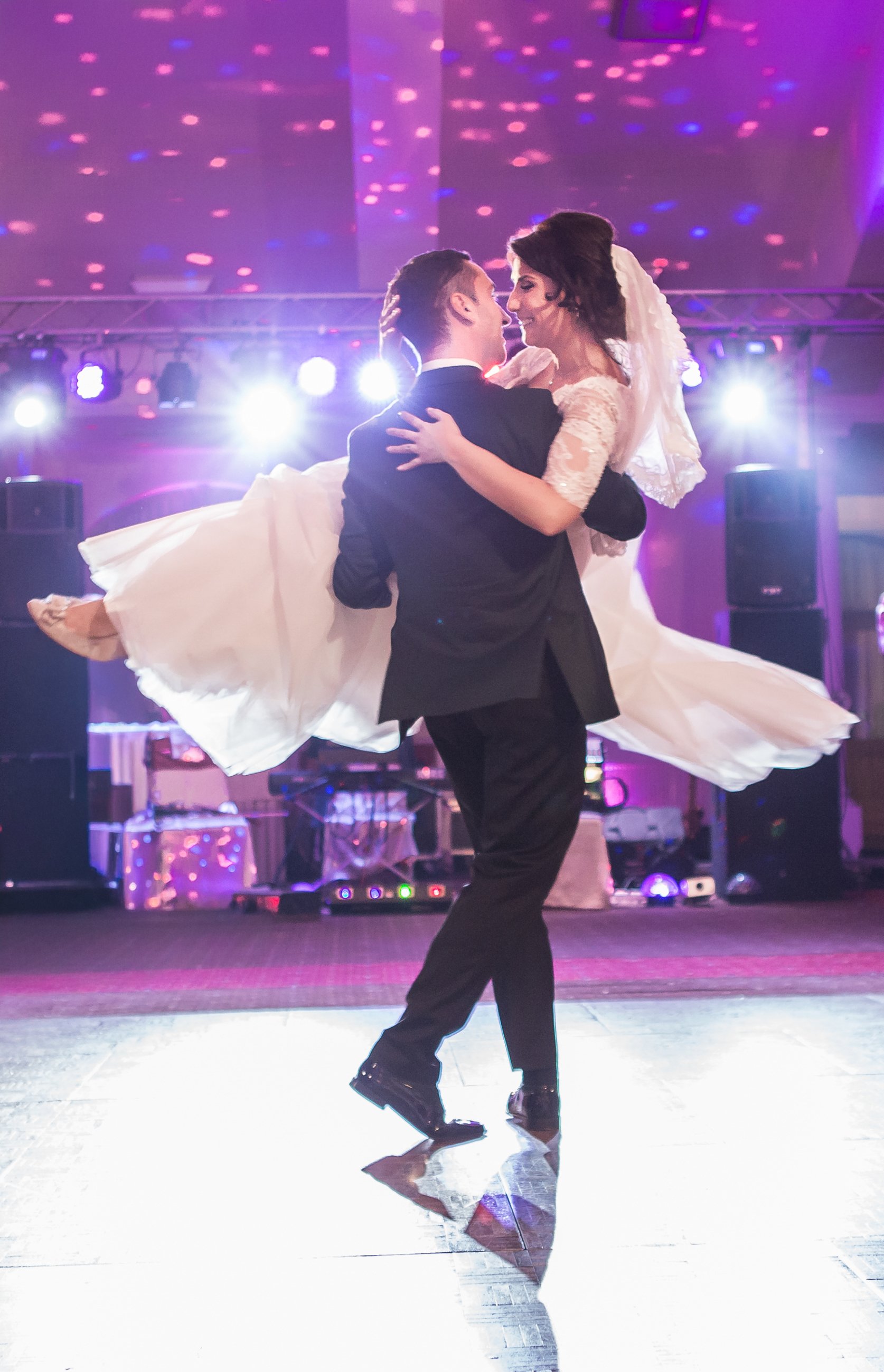 A couple dances closely at a formal event with purple lighting. The woman wears a white dress with lace sleeves and a veil, while the man wears a black suit. The background has bright stage lights and speakers.