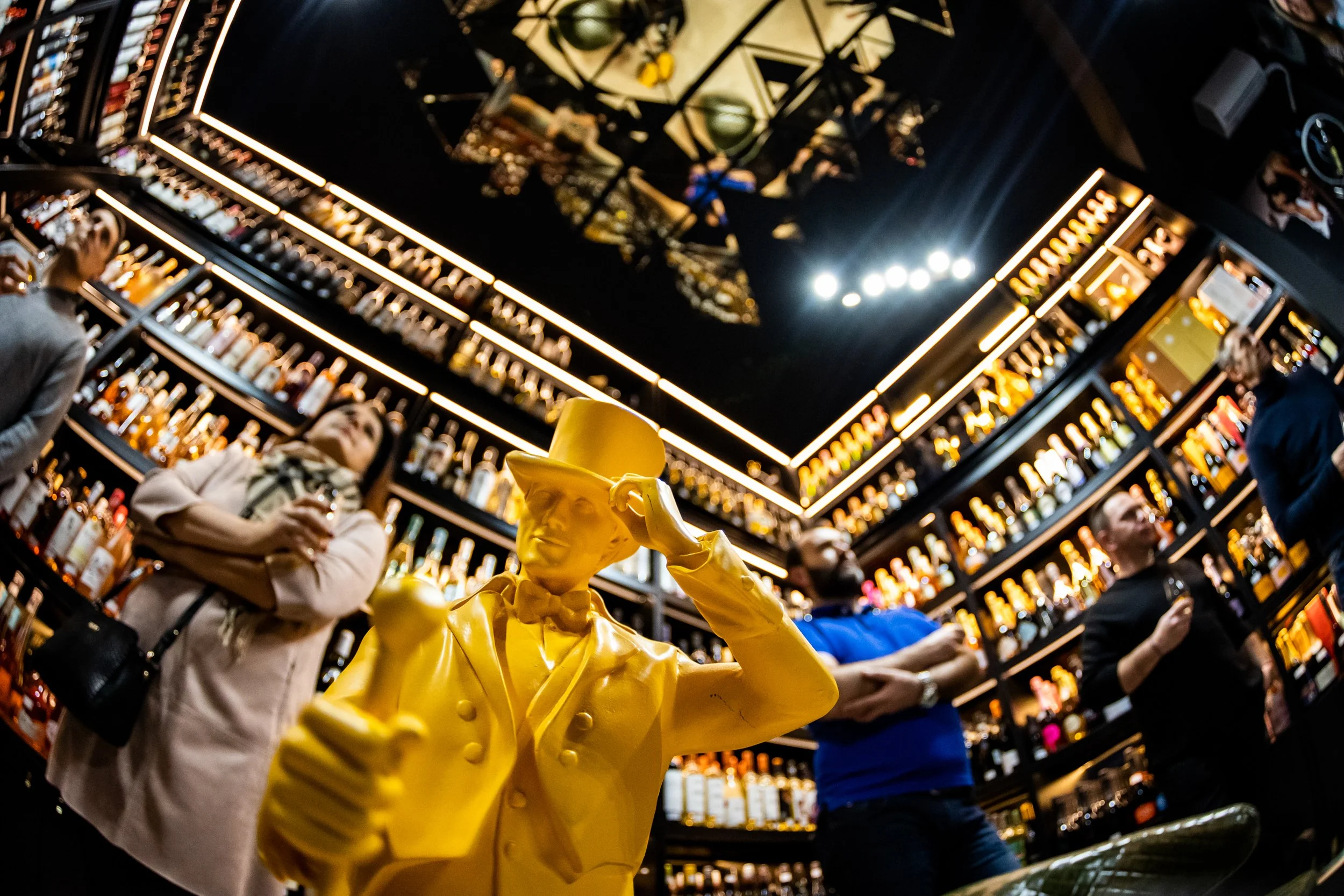 A yellow statue of a man wearing a hat, positioned in the foreground of a bar or liquor store with shelves of various bottles. Shoppers are in the background, and the ceiling has decorative black and gold patterns with lighting.