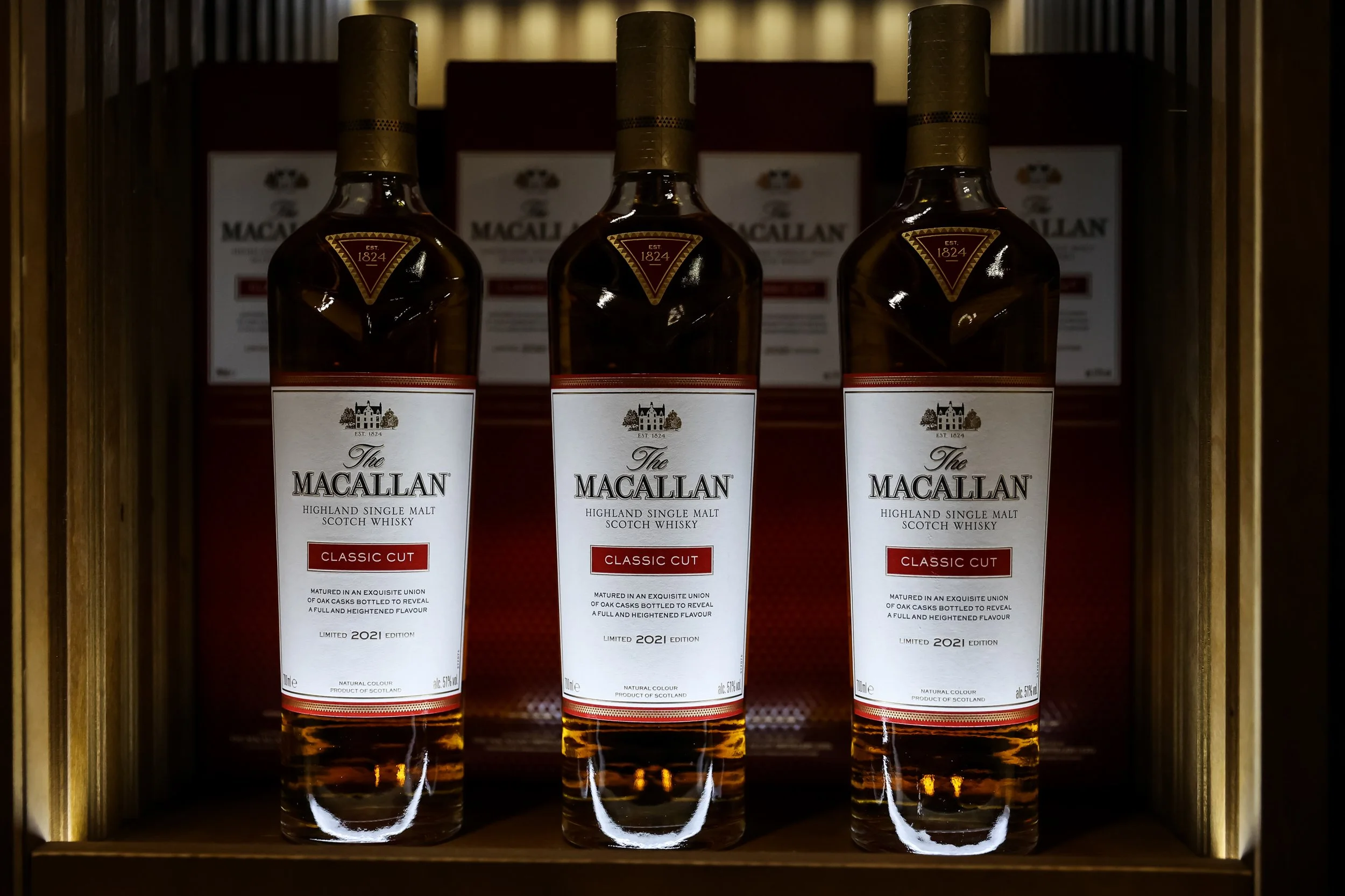 Three bottles of The Macallan Highland Single Malt Scotch Whisky Classic Cut on a wooden shelf, with a red and white label, in front of boxes with the brand name.