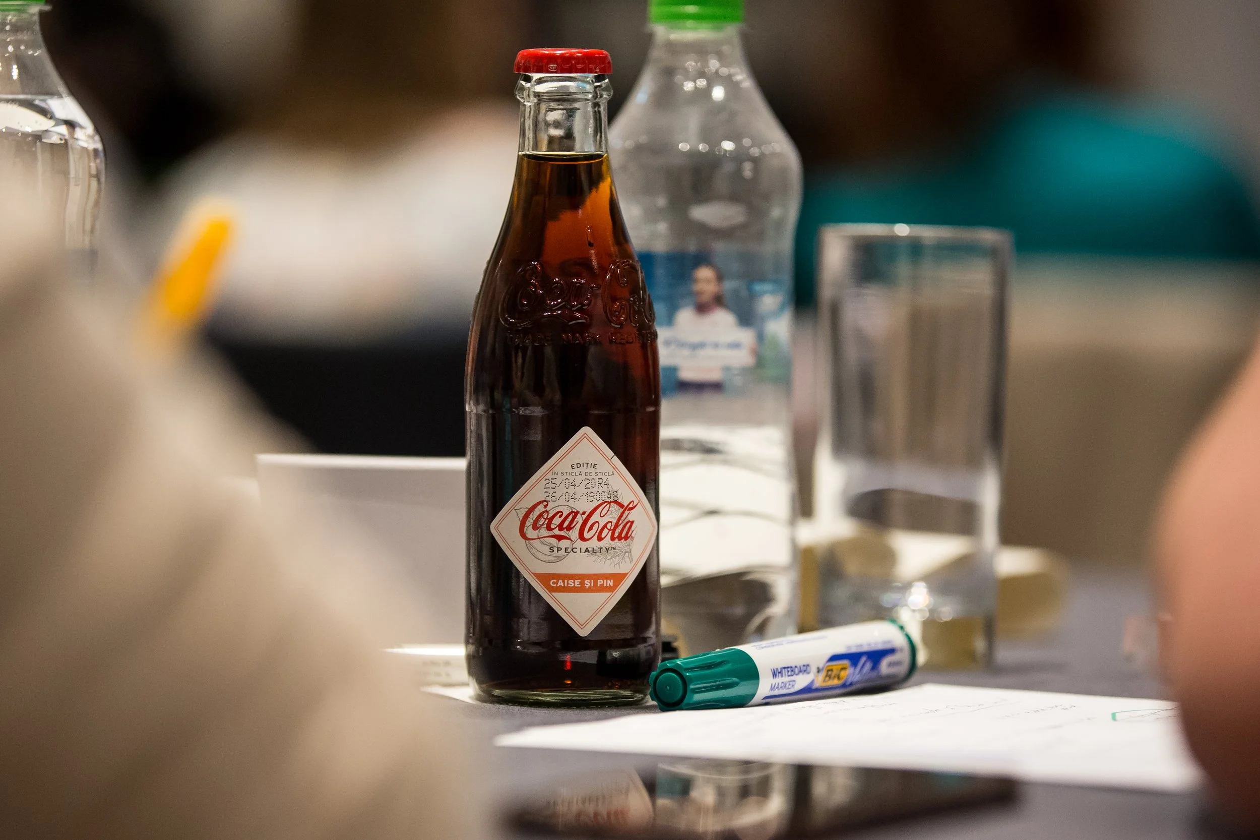A glass Coca-Cola bottle with a red cap, placed on a table with various objects including a water bottle, a pen, and a glass. The background is blurred.