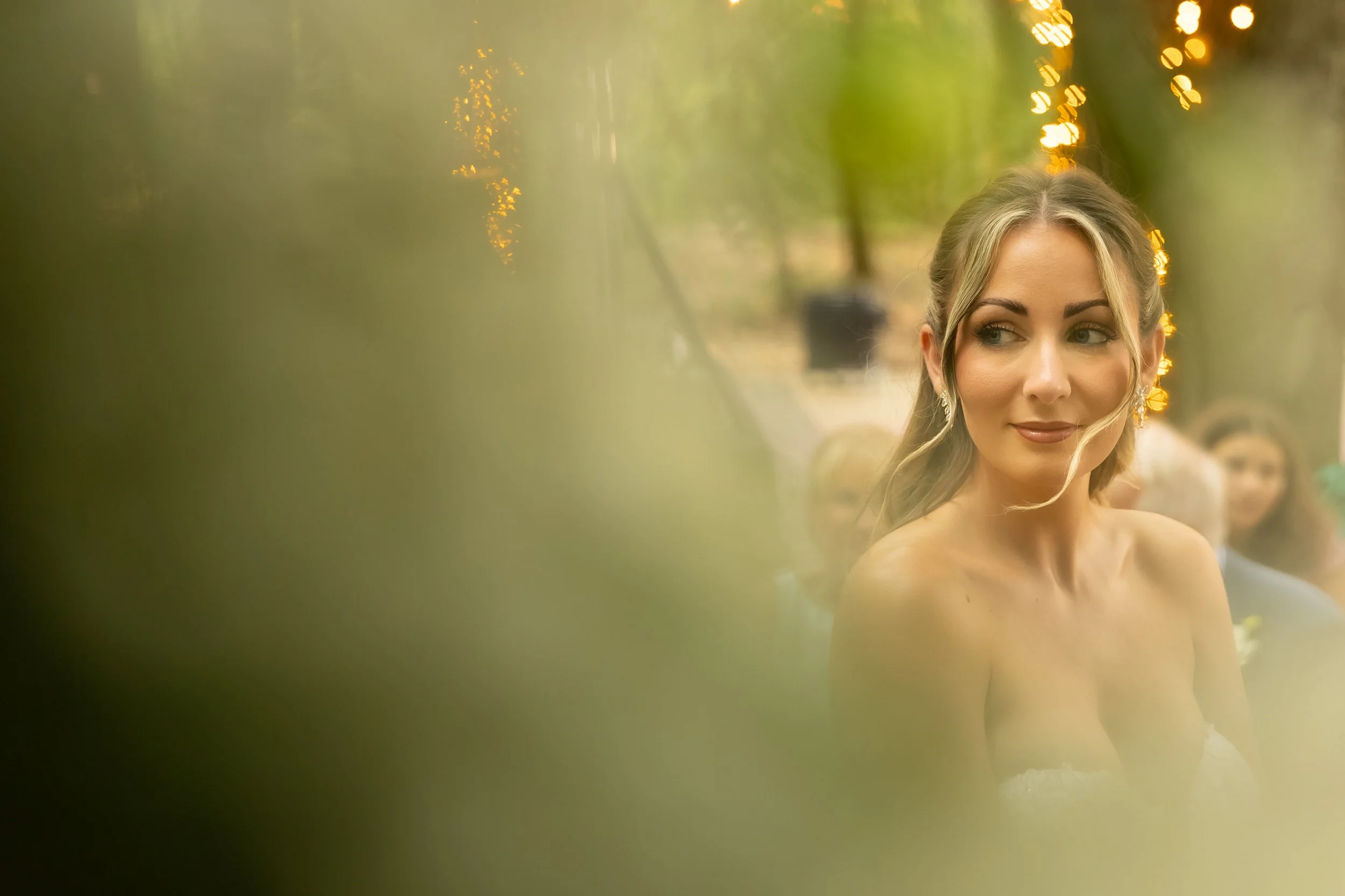 A woman with blonde hair and earrings, smiling softly, outdoors during the evening with string lights in the background.