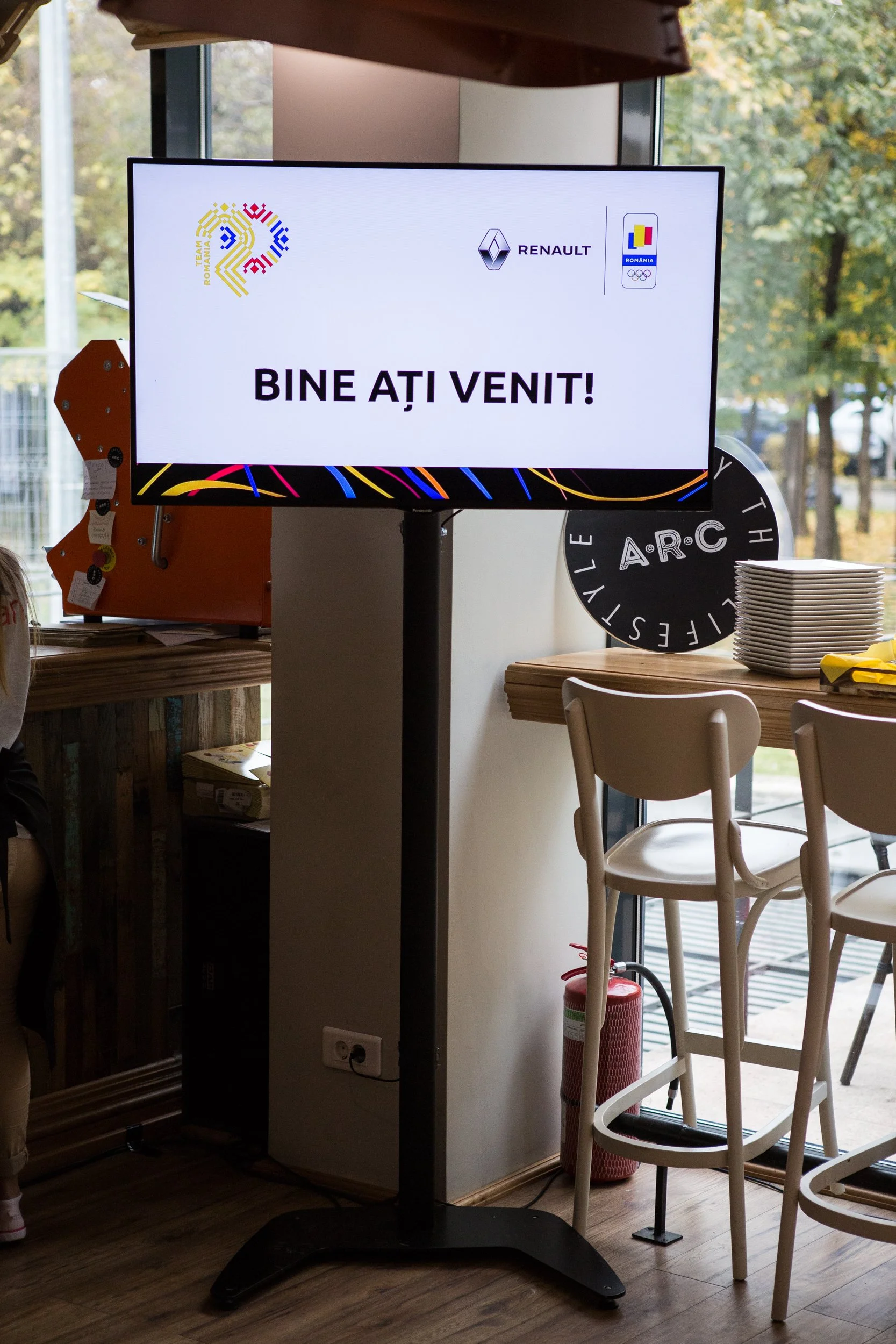 An indoor scene with a large digital screen displaying a message in Romanian, surrounded by chairs and a table. The screen has logos of Renault, Team Romania, and Romania's Olympic team. The message on the screen says 'BINE AȚI VENIT!', meaning 'WELC