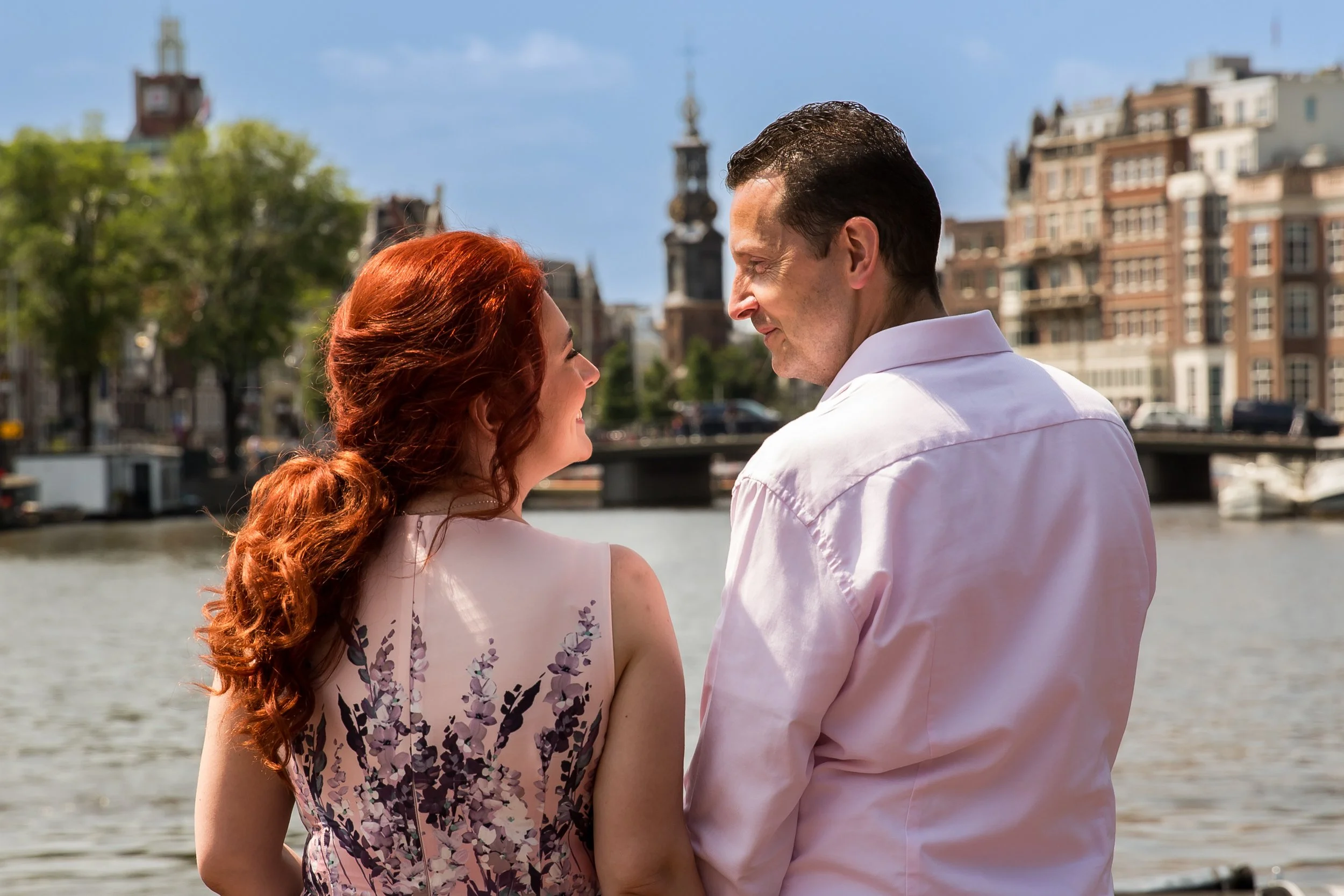 A man and a woman are standing close together near a body of water, smiling and looking into each other's eyes, with buildings and a monument in the background.