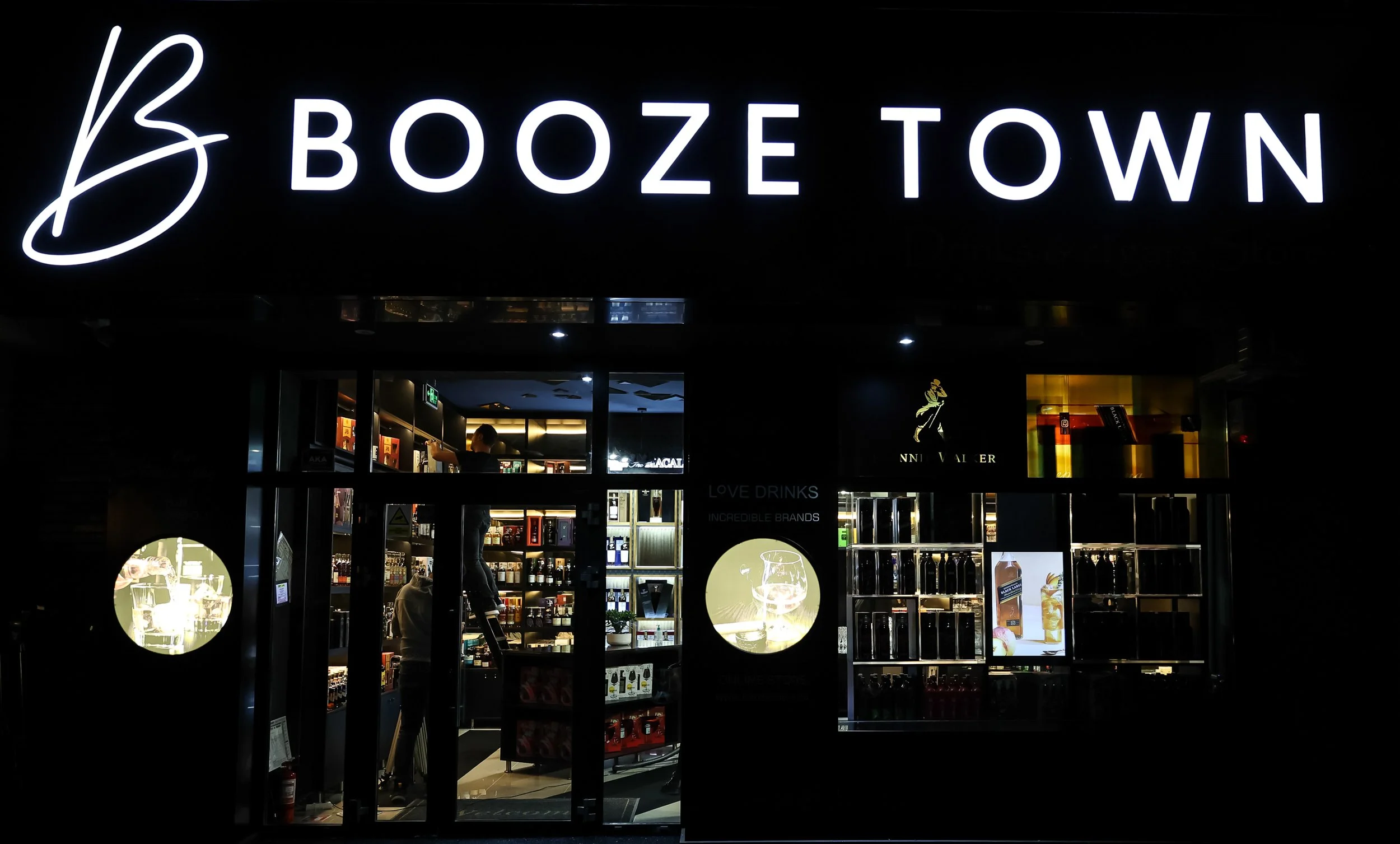 Night view of a store entrance with illuminated sign reading 'Boozetown'; inside the store, shelves with various bottles and products visible through large windows.