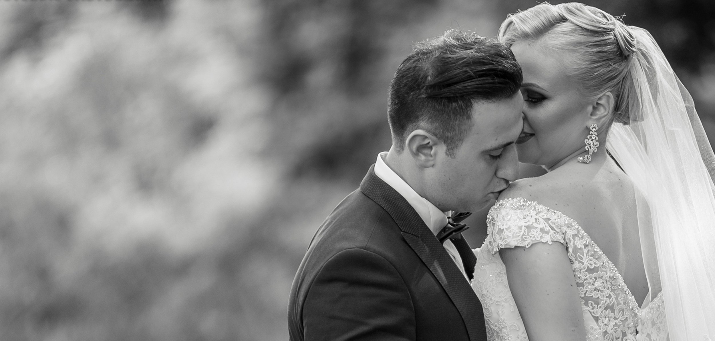 A black and white photo of a bride and groom kissing during their wedding. The groom is wearing a tuxedo, and the bride is in a lace wedding dress with earrings and a veil.