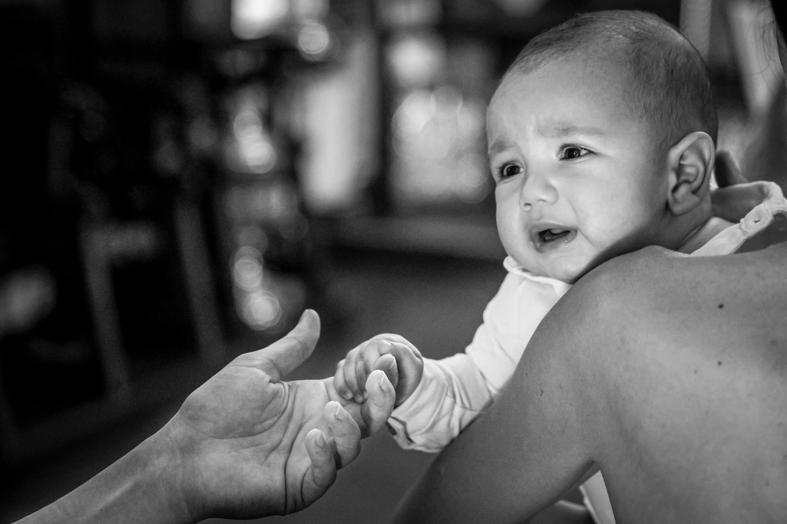 A black and white photo of a baby with a shaved head and big eyes, smiling and reaching out to hold someone's hand, with a blurred background.