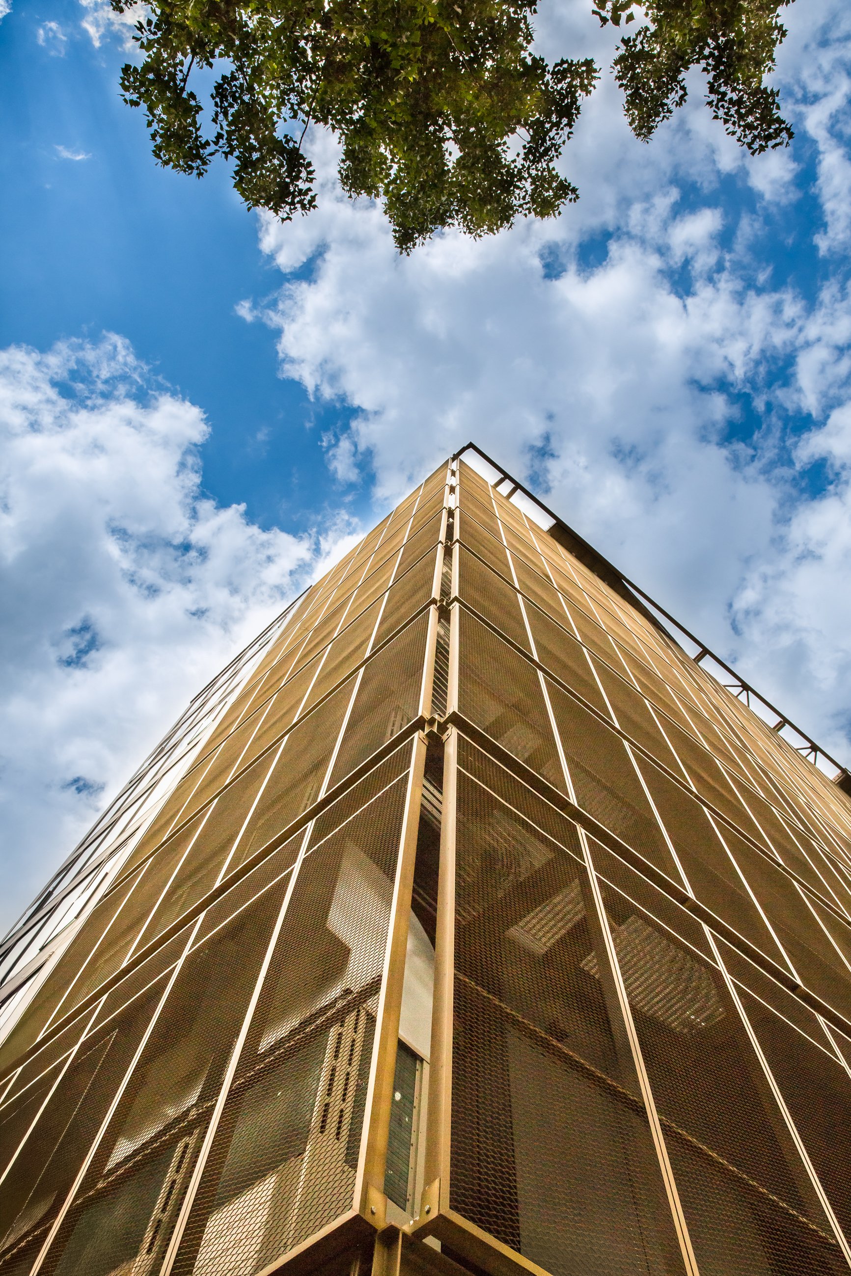 A tall modern building with a glass and metal facade viewed from the ground looking up toward the blue sky with scattered clouds.