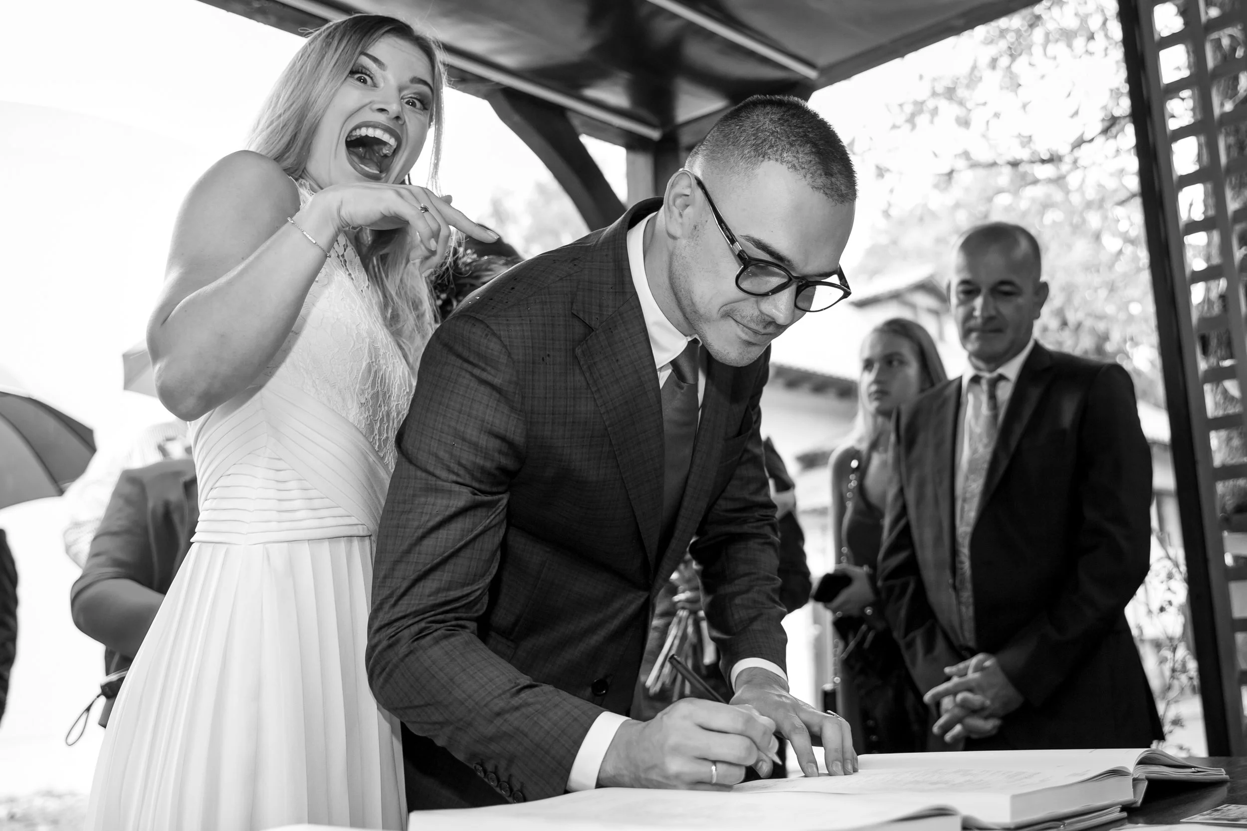 A woman in a wedding dress is excitedly pointing and smiling at a man in a suit who is signing a document at an outdoor event.