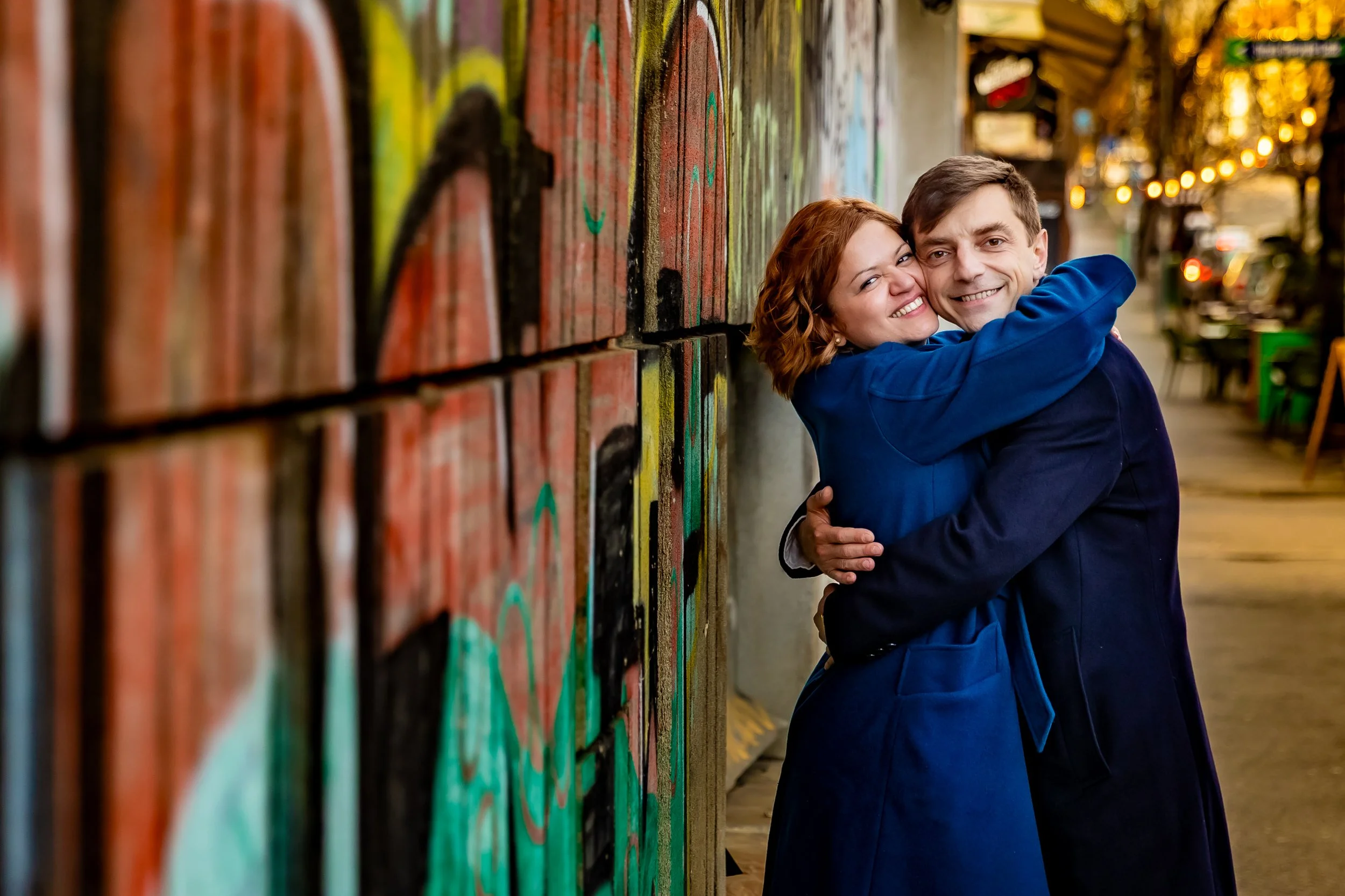 A young couple hugging and smiling against a colorful brick wall with graffiti art, in an outdoor urban setting with warm lighting and blurred background.