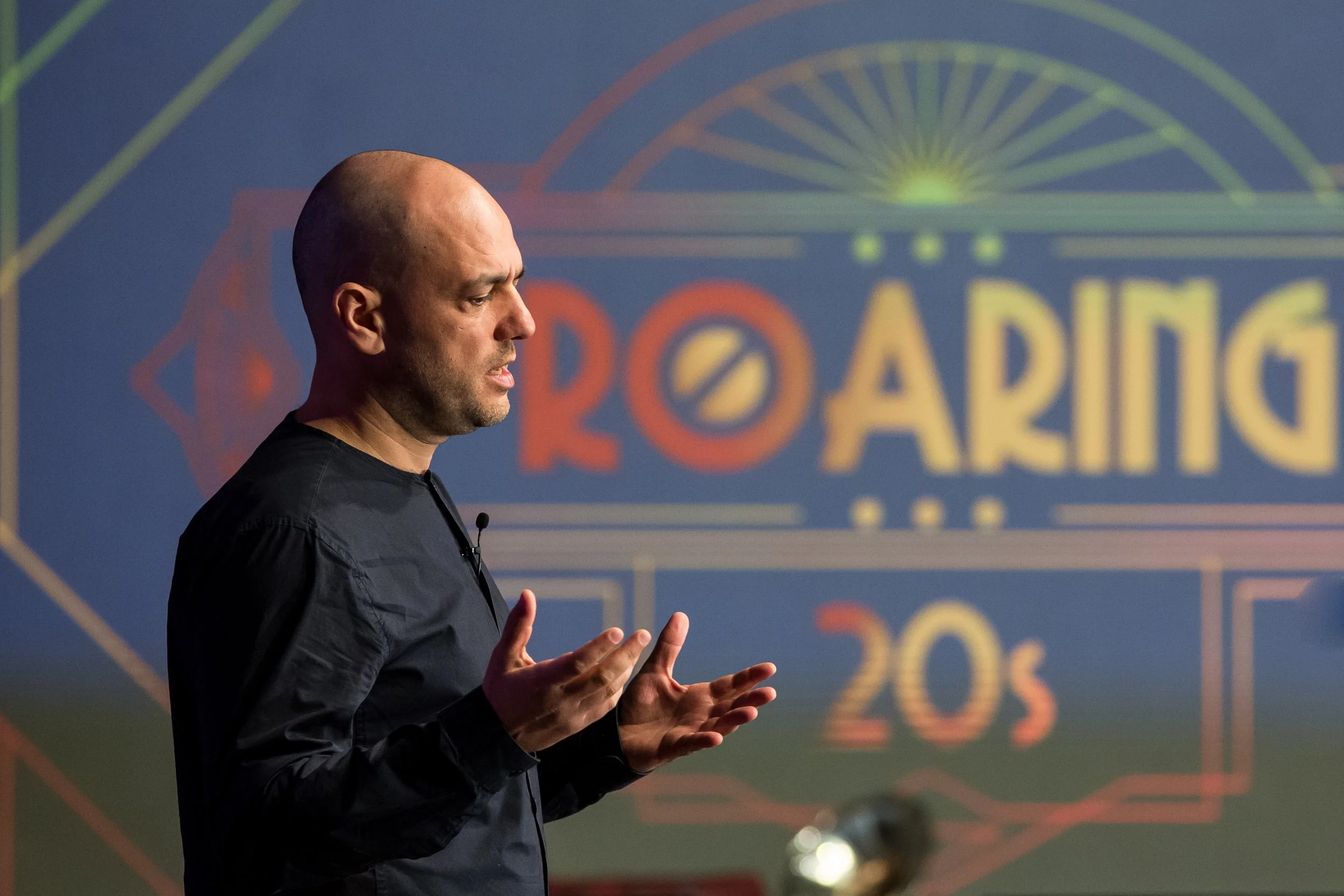 A man with a shaved head and wearing a black shirt is speaking or presenting, with a colorful background featuring the word 'PARIS' and a stylized Ferris wheel design.