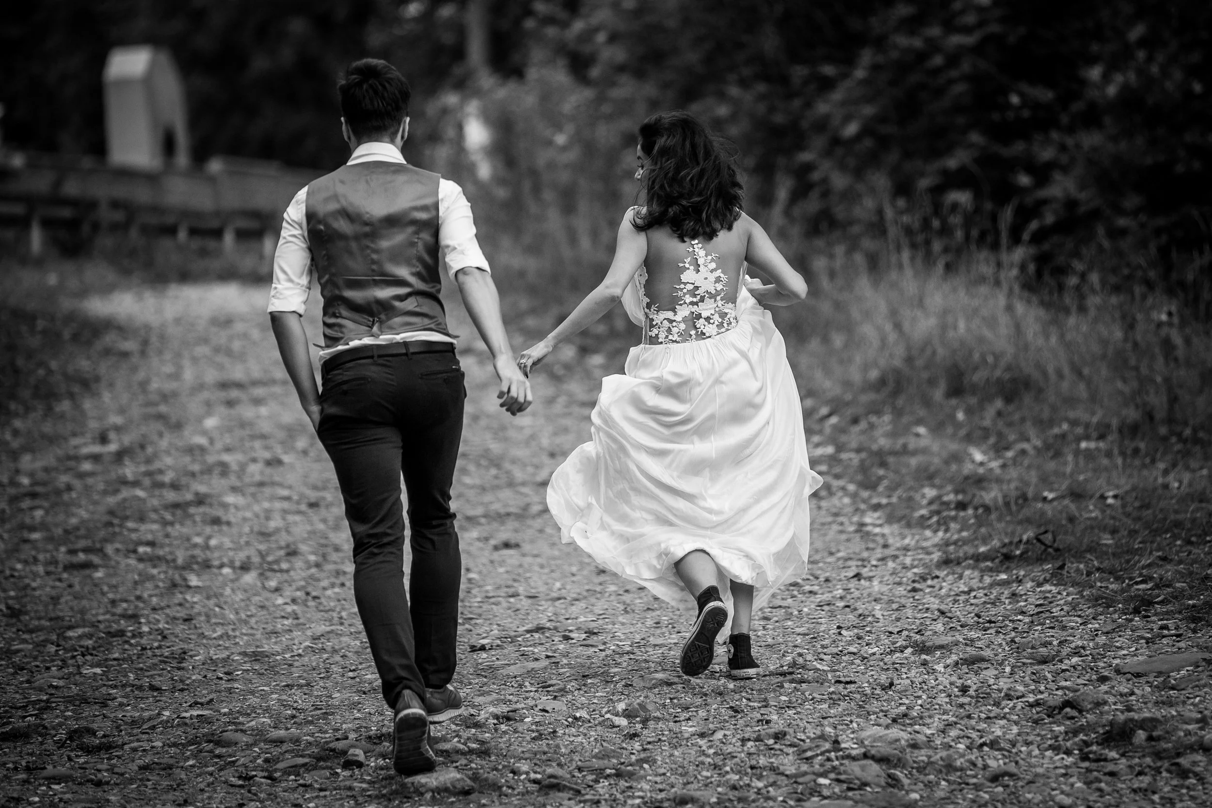 A black and white photo of a couple running hand in hand on a dirt path in nature. The woman wears a wedding dress with floral lace details on the back, and the man is dressed in a vest and dress shirt.