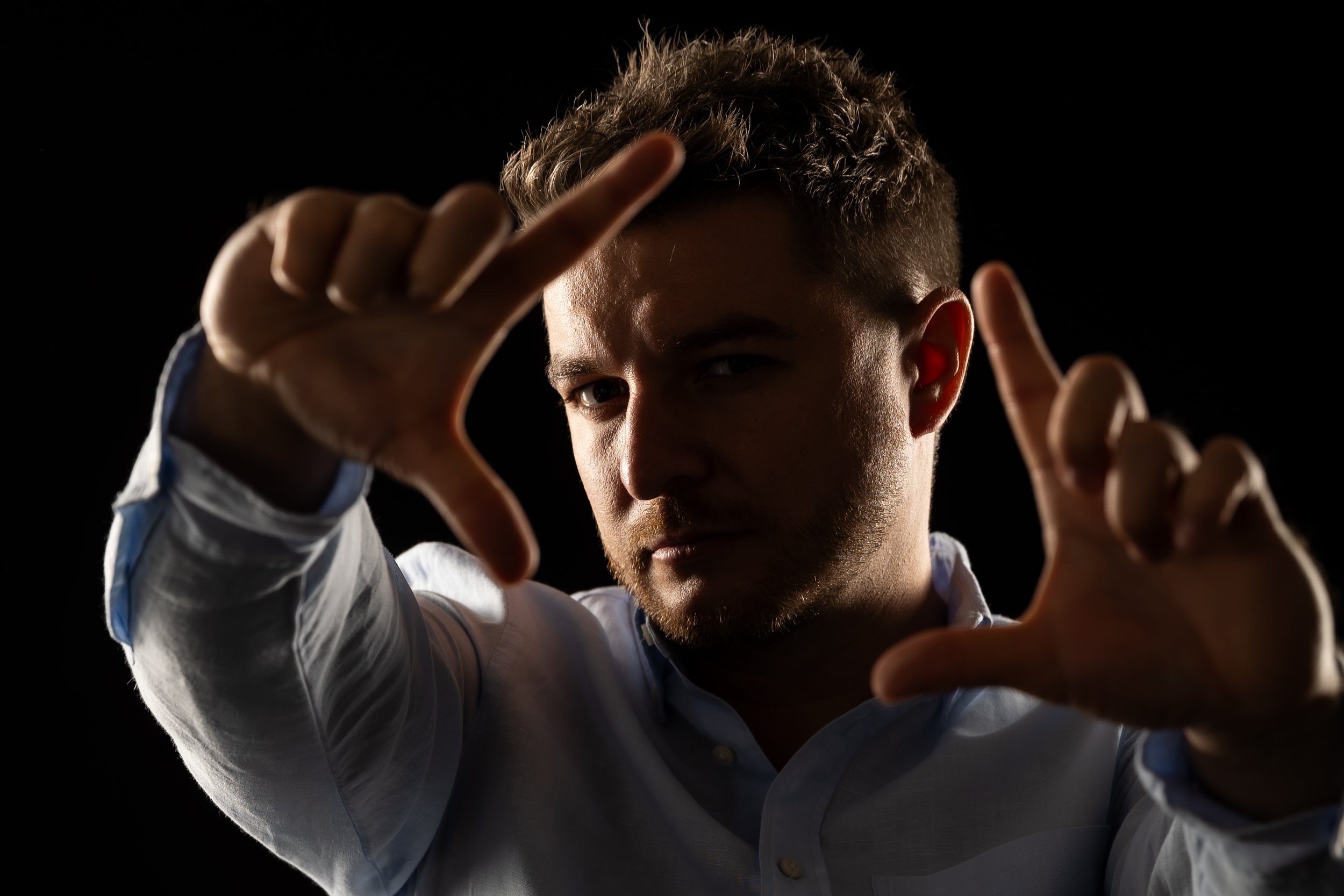 A young man with short curly hair and a light beard, looking directly at the camera, making a framing gesture with his hands against a black background.