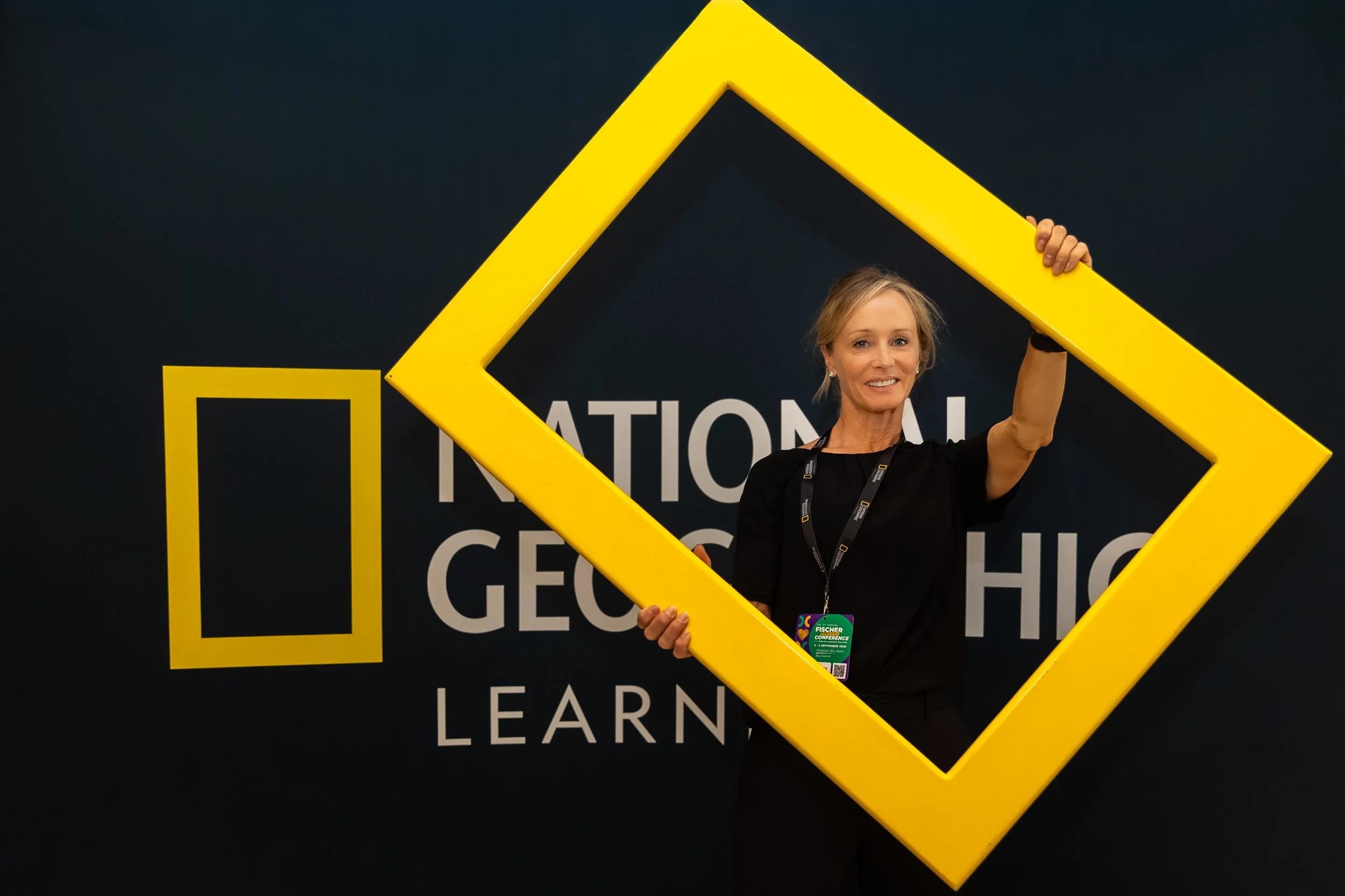A smiling woman holding a large yellow frame at a conference with a dark background that has the words 'NATIONAL GEOGRAPHIC' and 'LEARNING' visible.