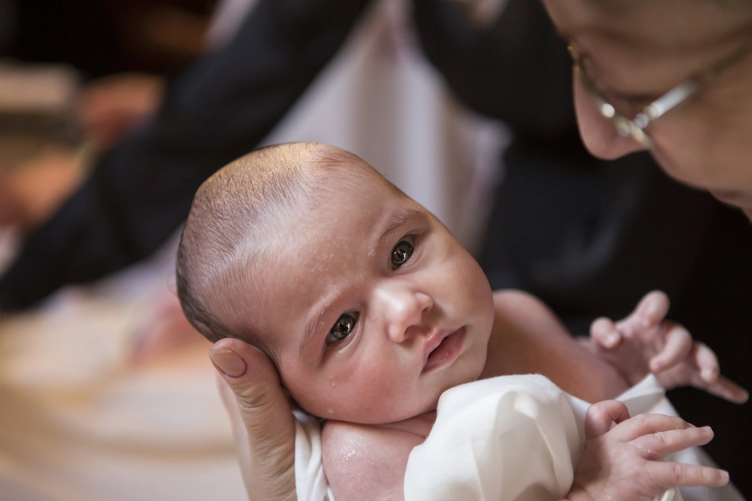 Close-up of a baby with big eyes lying on its back, being gently held and supported by an adult's hand, with the adult's face leaning close to the baby's face.