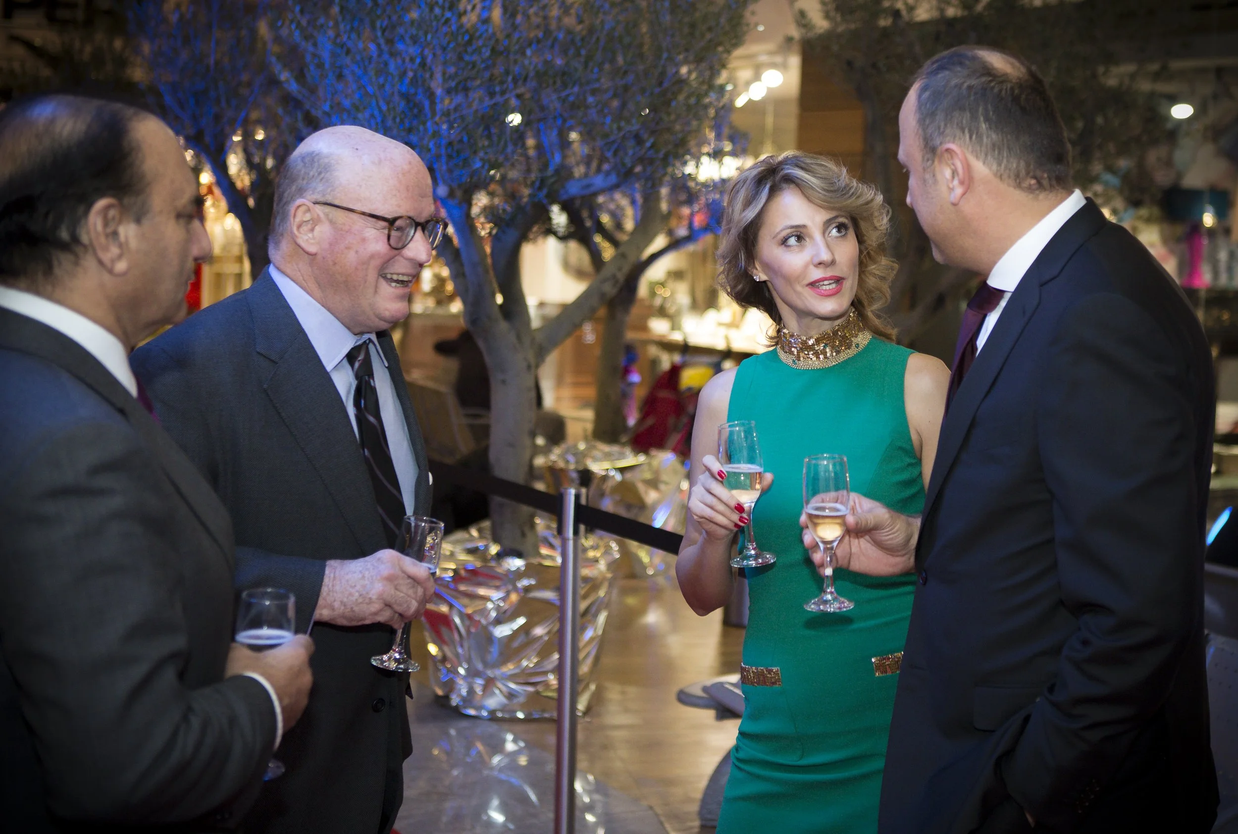 A group of people dressed in formal attire, holding glasses of wine, engaged in conversation at an indoor event with a tree decorated with blue lights in the background.