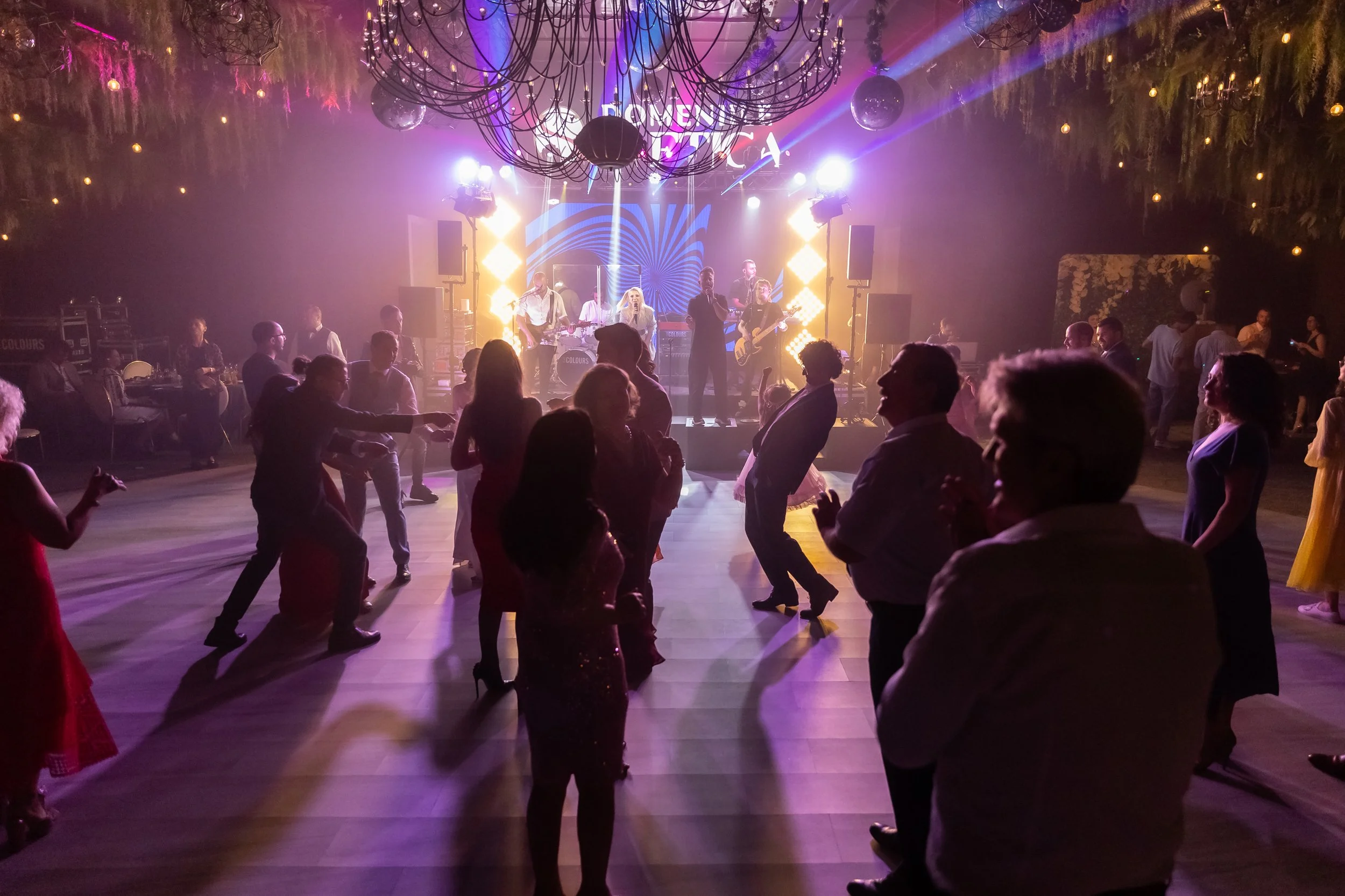 People dancing and enjoying live music at an indoor or outdoor event with stage lighting, a band performing on stage, and decorative hanging lights and greenery.