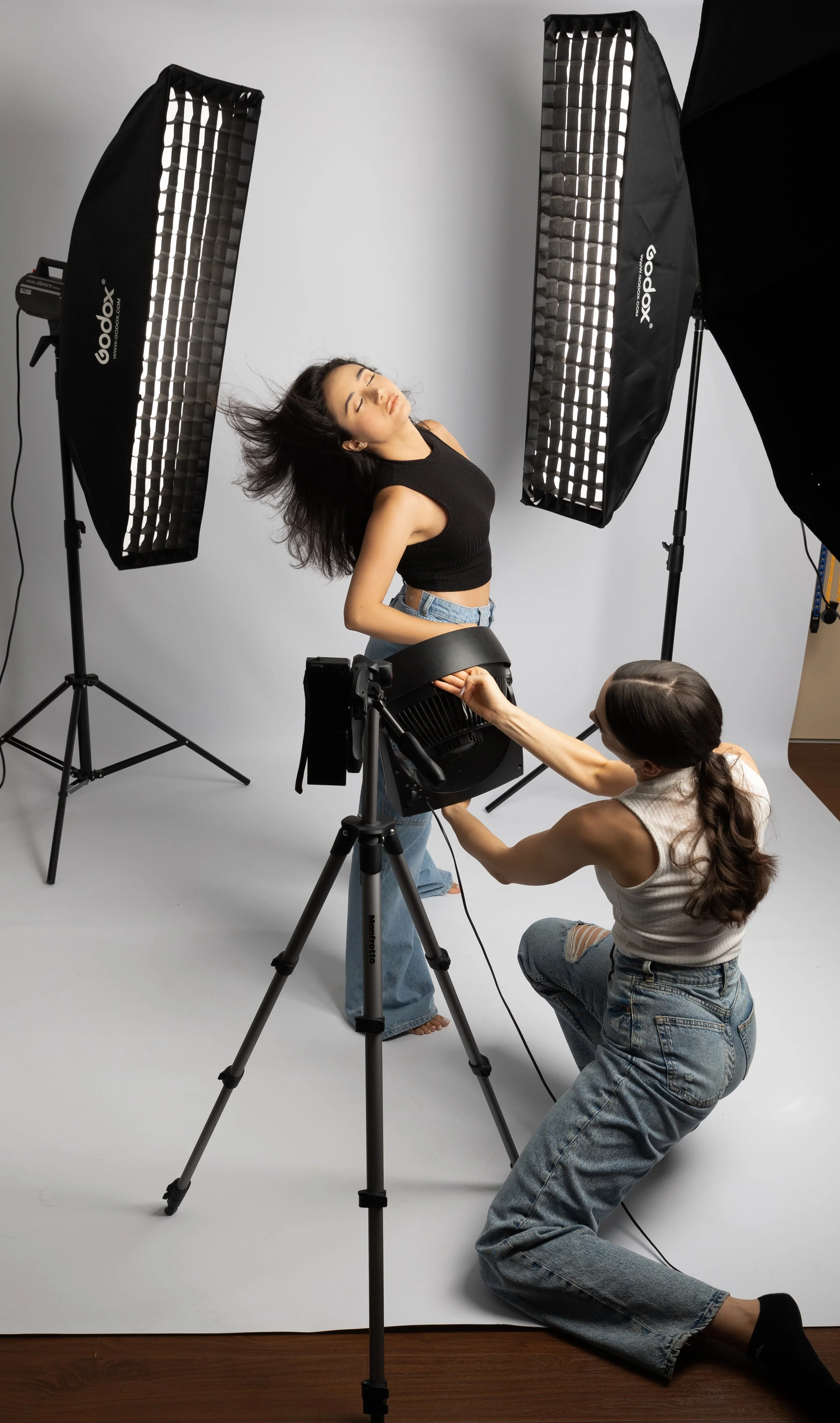 A woman posing with her eyes closed and hair blowing, while another woman adjusts lighting equipment in a photography studio with large studio lights and a white backdrop.