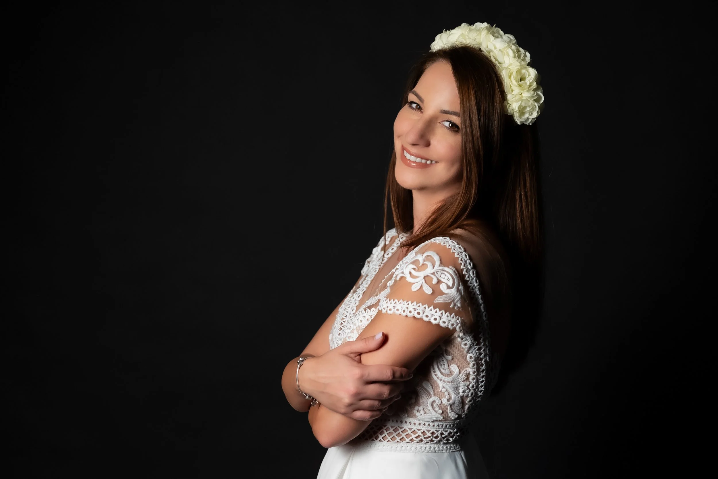 A smiling woman with long brown hair wearing a white lace dress and a floral headband, standing against a black background.