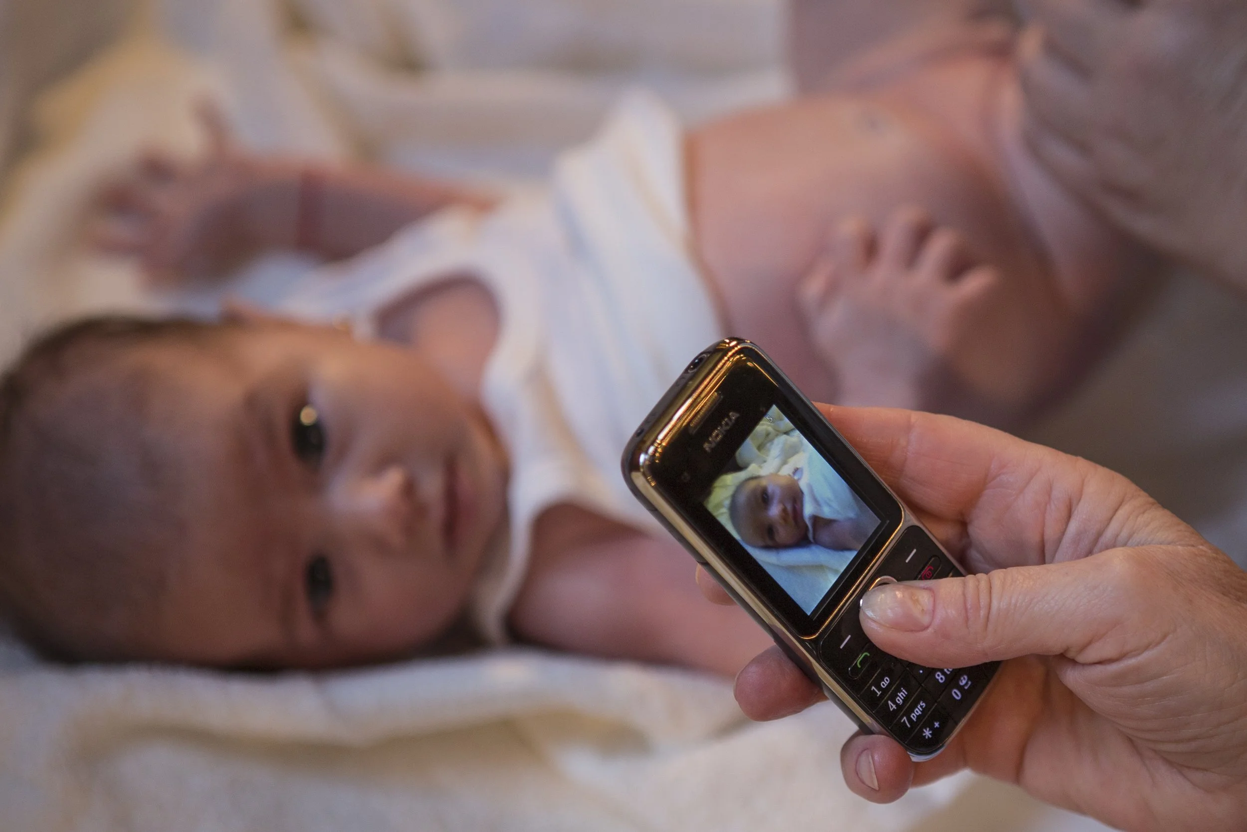 A person holding a flip phone with a photo of a baby displayed on the screen, lying on a surface, with another person next to the baby.