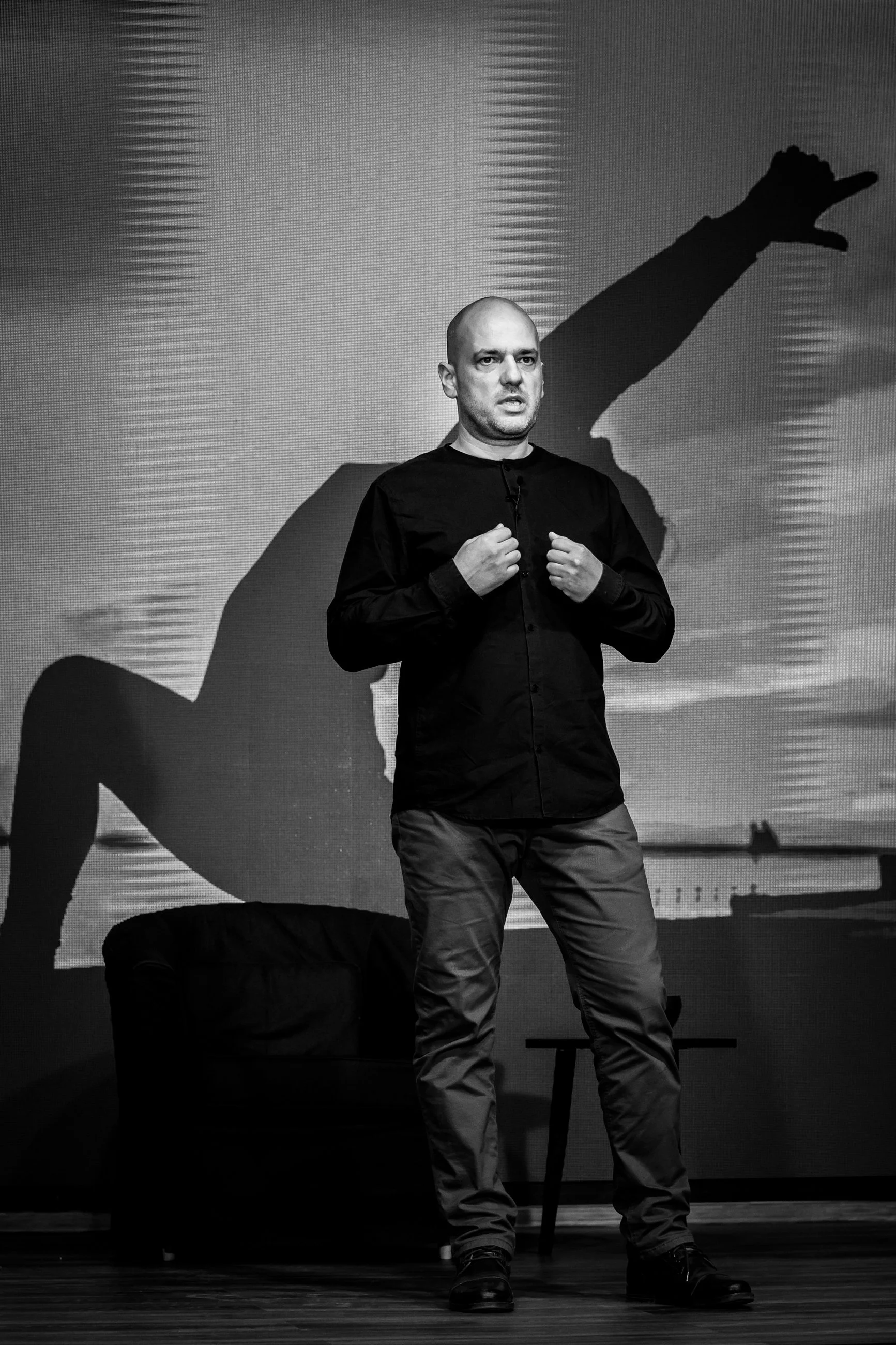 Black and white photo of a man with a shaved head giving a presentation on stage. He is wearing a dark shirt and gray pants. Behind him, a large screen displays a silhouette of a person running along a shoreline.