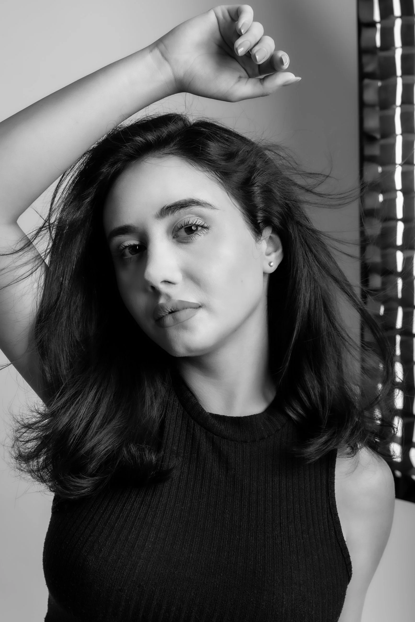 Black and white photo of a young woman with long wavy hair posing with her arm raised above her head, wearing a sleeveless top, facing slightly to the side, in front of a neutral background.