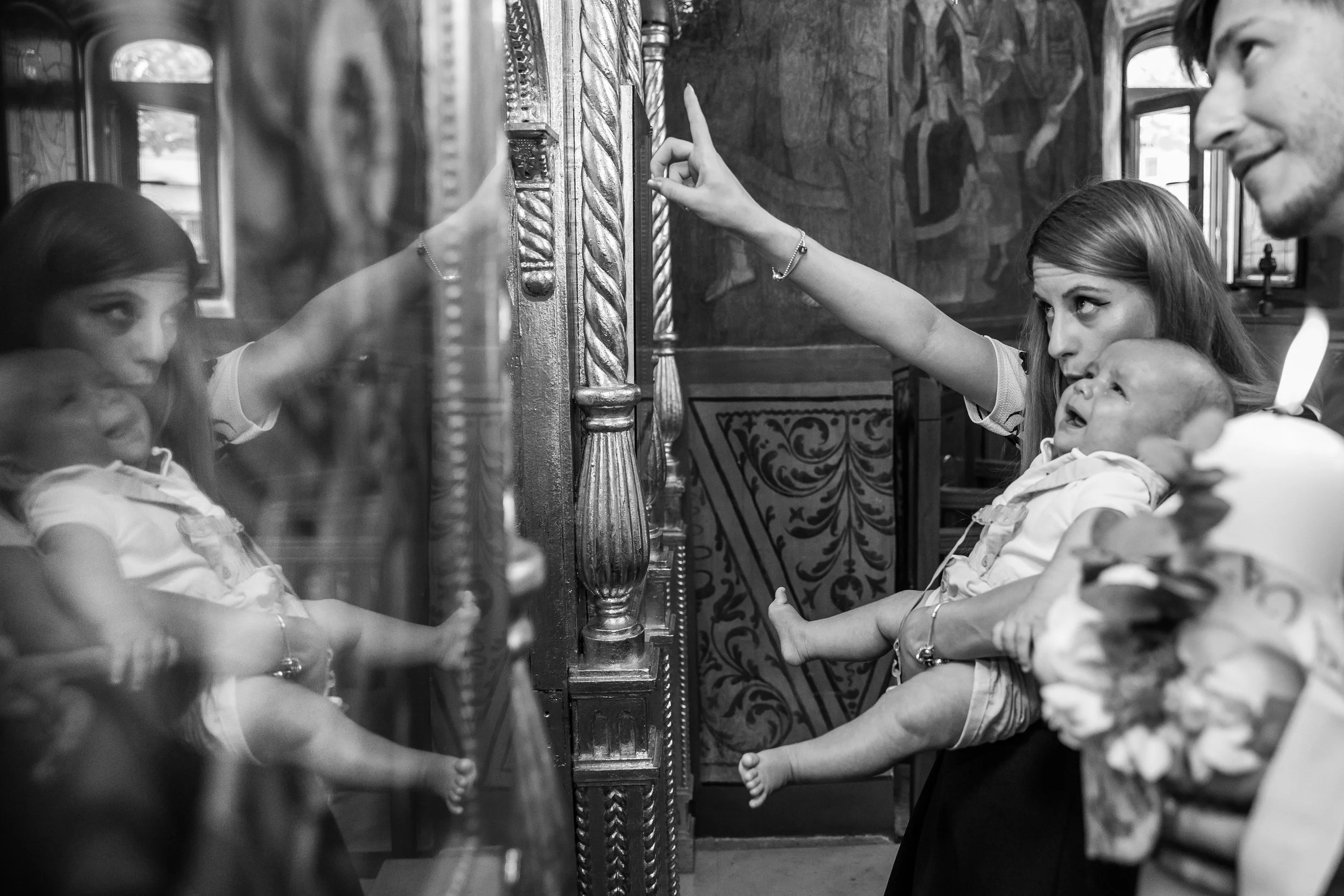A woman is holding a baby while looking at a young girl inside a church. The girl is reaching out to touch an ornate, carved wooden stand with religious icons. A man is smiling in the background.