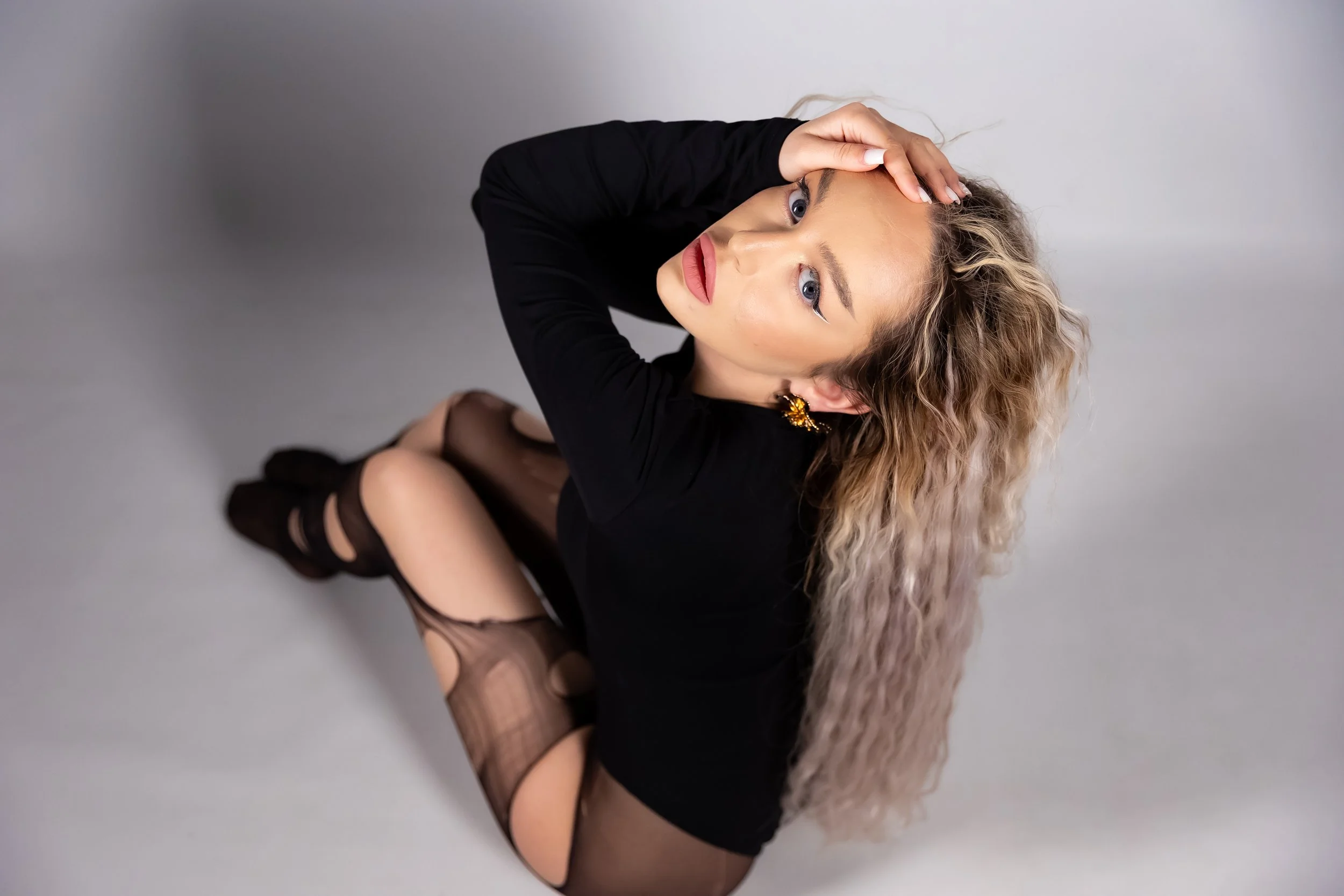 A woman with curly blonde hair, blue eyes, wearing black clothing, fishnet stockings, and black high heels, sitting on the floor against a plain light gray background.