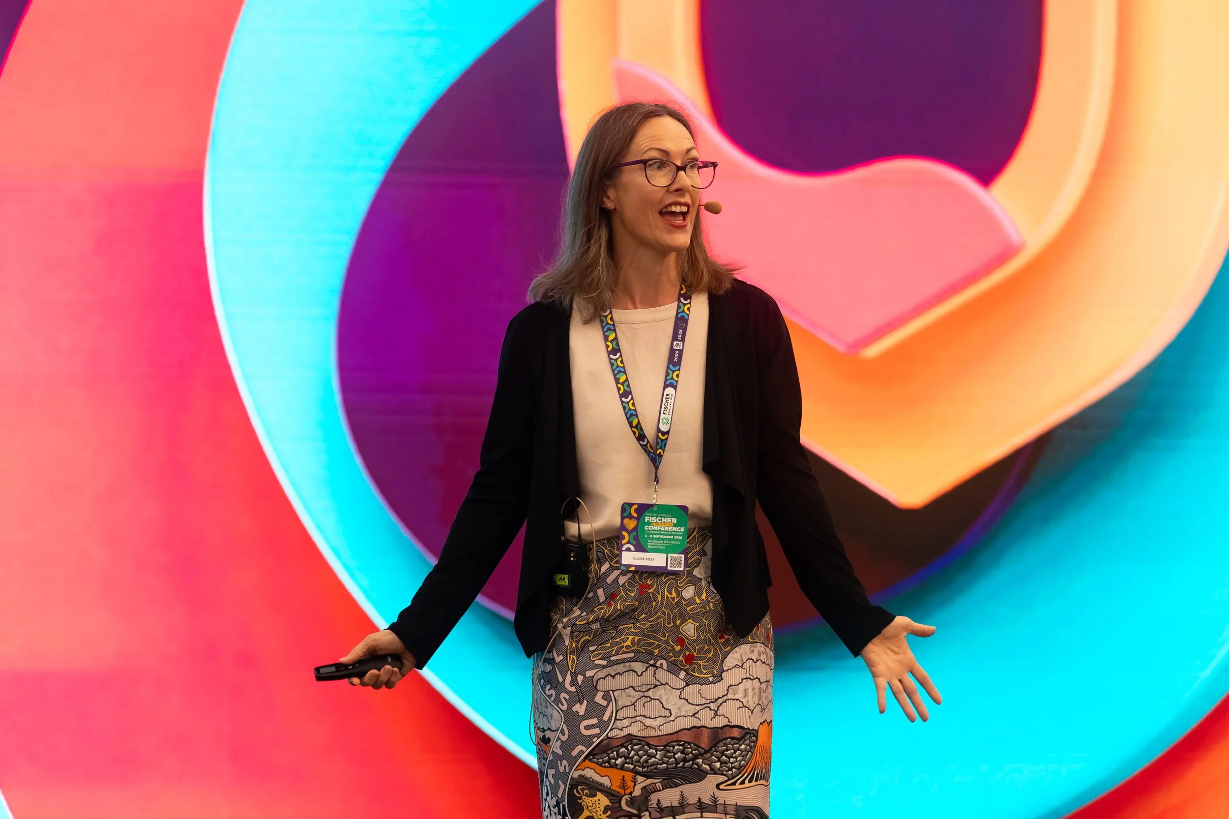 A woman speaking at a conference, gesturing with her hands, standing in front of a colorful, abstract background. She has glasses, a microphone headset, and a conference badge around her neck.