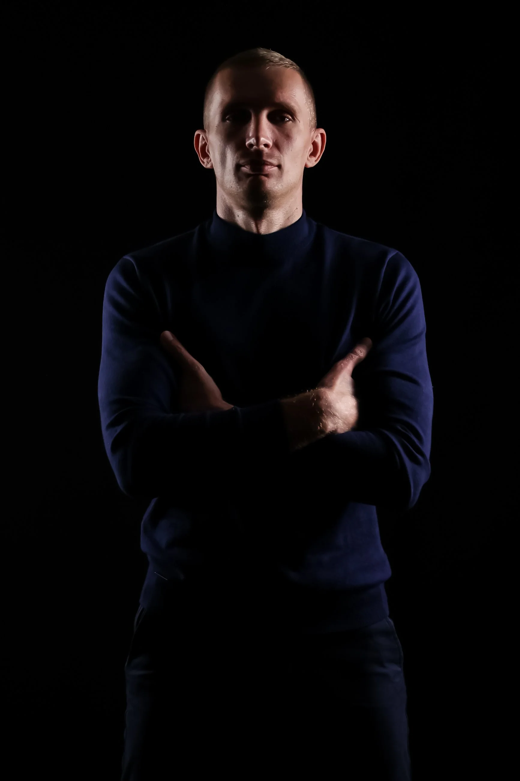 A man standing with arms crossed against a dark background, wearing a dark long-sleeve shirt, with a serious expression.