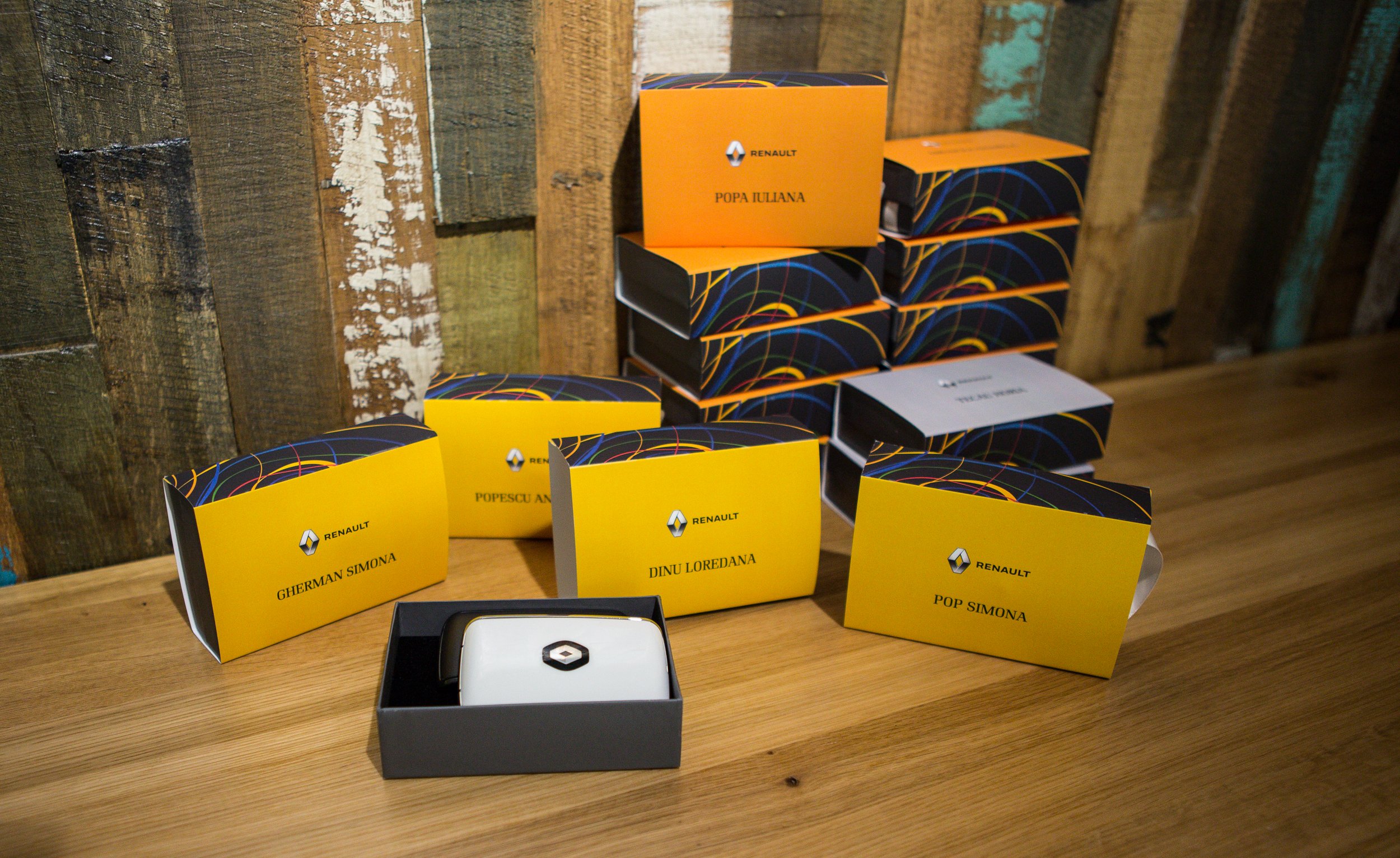 Several yellow and black boxes with the Renault logo on a wooden table, some labeled with nicknames and names. A white square device with the Renault logo is in a black box in the foreground. The background features a rustic wooden wall.