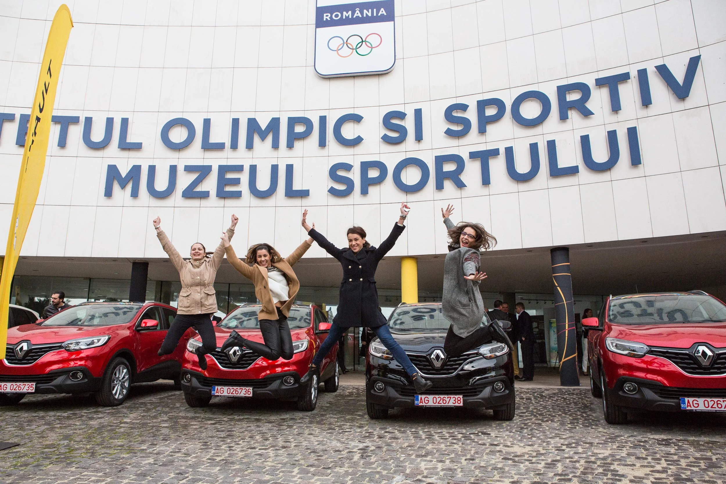 Four women jumping and smiling in front of a car dealership with a large sign that reads 'Muzeul Sportului' and an Olympic Romania emblem. The background shows several red and black cars with Romanian license plates, and a building with a sign in Rom