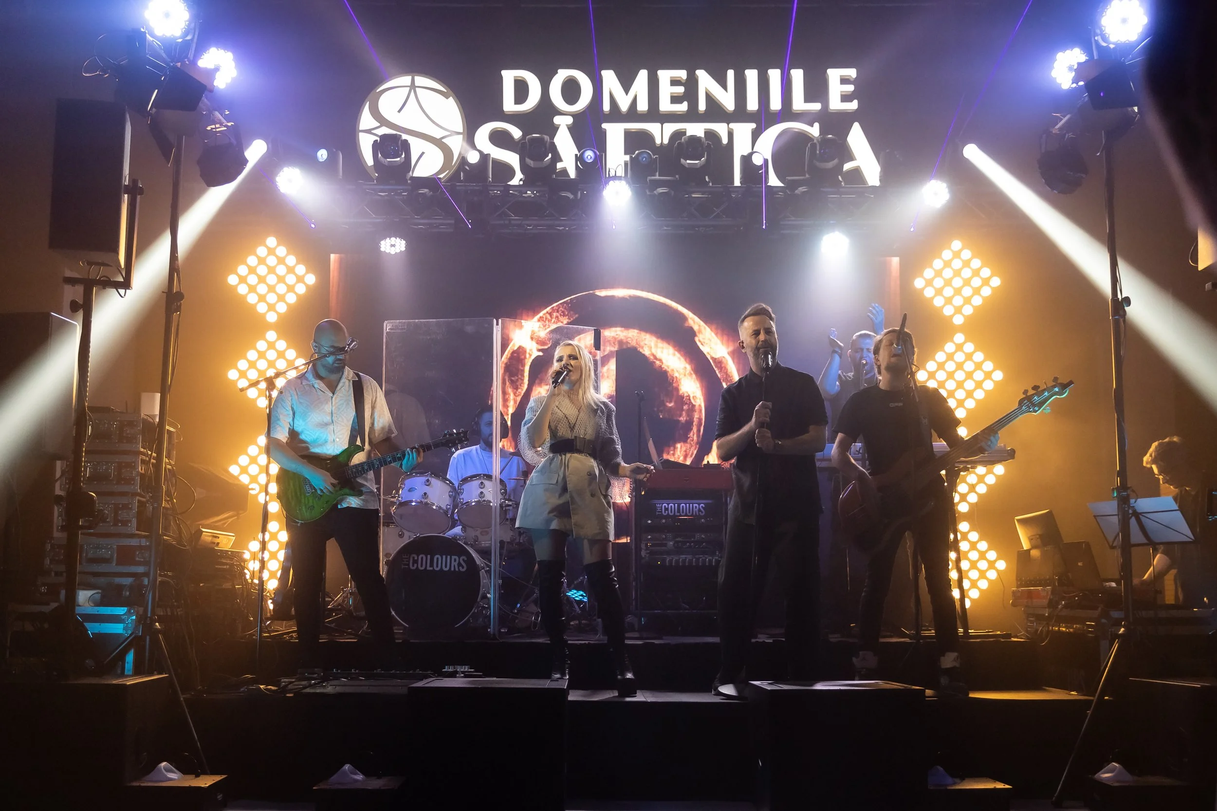 A live music performance at the Donnille Saefica event, with band members playing instruments and singing on stage, illuminated by colorful stage lights and background graphics.