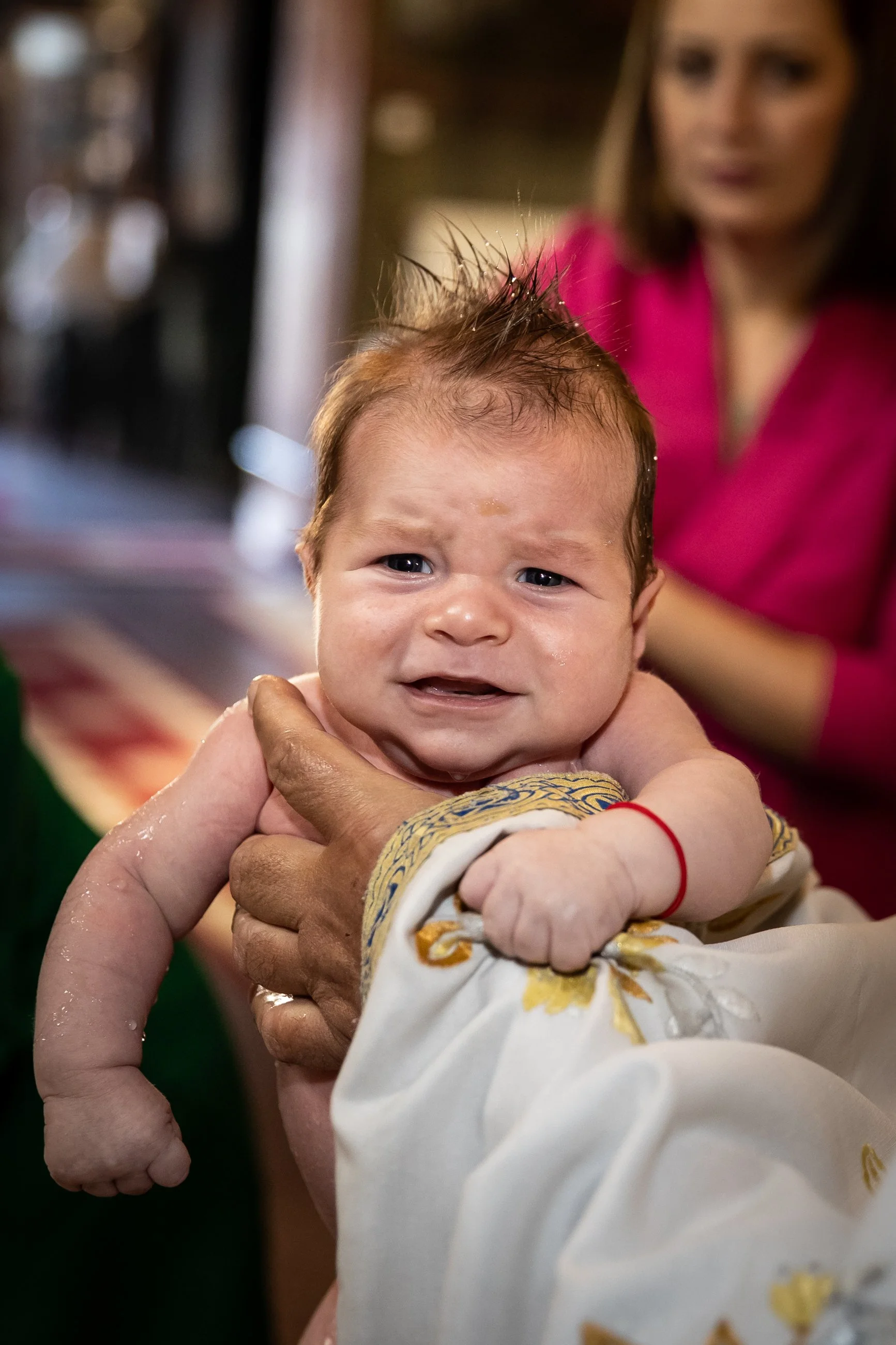 A baby with wet hair being held by an adult, with a woman in a pink shirt in the background.