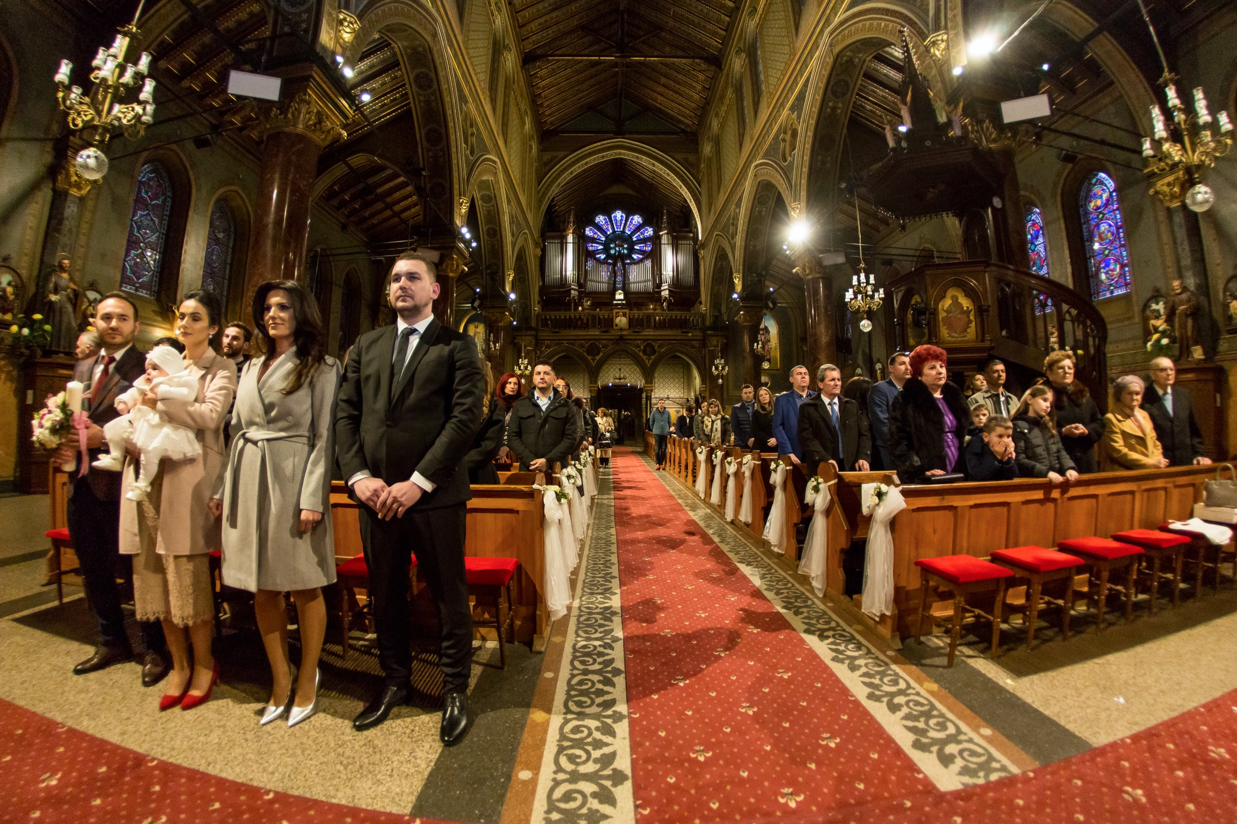 People standing inside a church, some dressed in formal attire, during a wedding ceremony or special event. The church features stained glass windows, ornate decorations, chandeliers, and a central aisle with a red carpet.