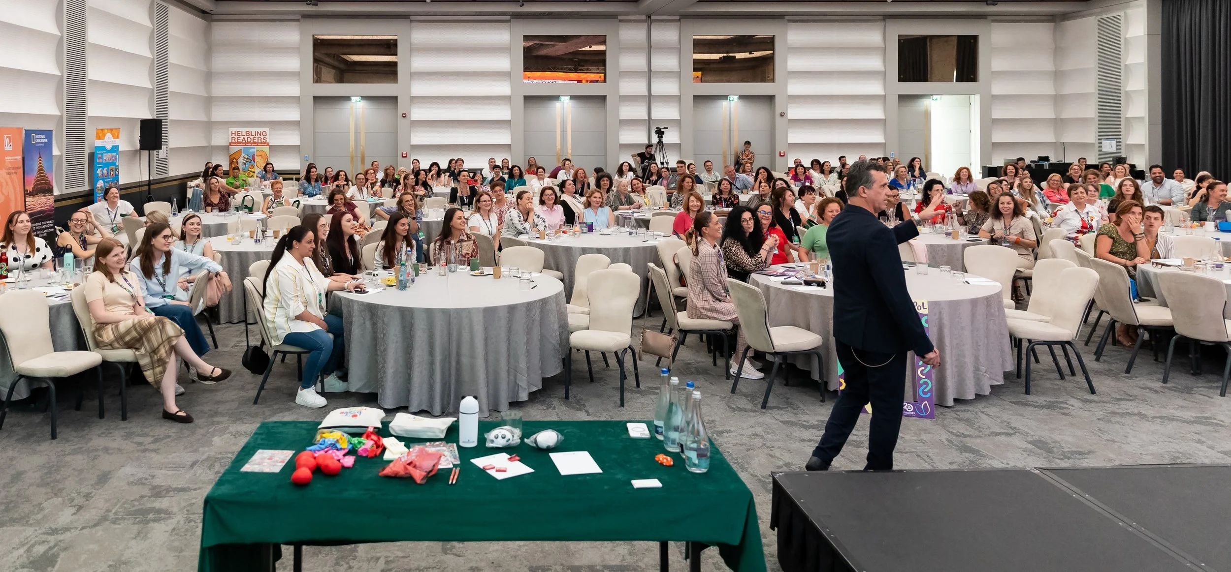 A large conference room filled with round tables and chairs, with many people attending a presentation or event. A man is speaking at the front, and there are banners and a camera setup in the background.