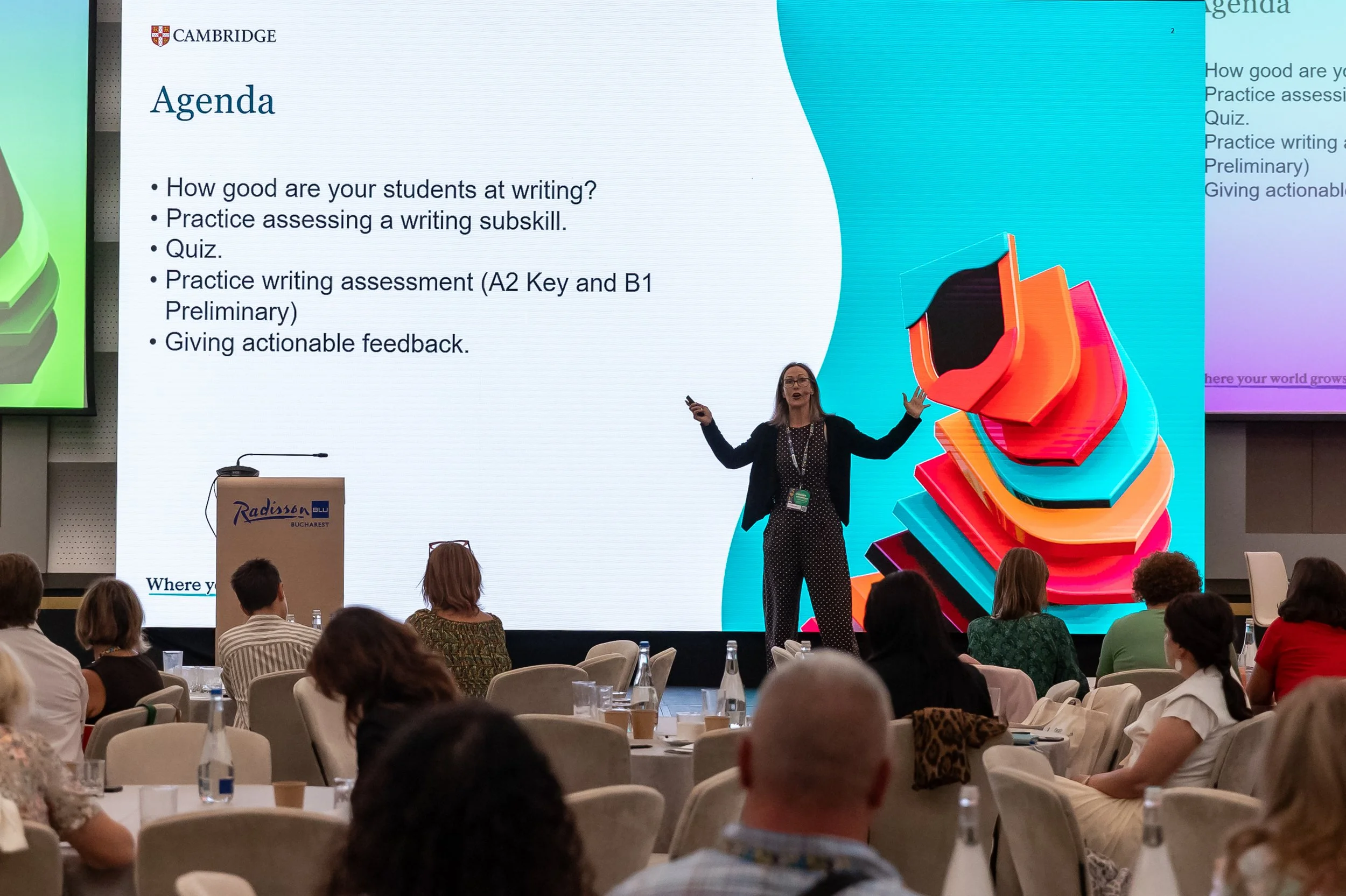 A woman is giving a presentation on a stage at a conference. The large screen behind her displays the agenda for a workshop, including topics like student writing assessment, quizzes, and feedback. Several audience members are seated in front of her,