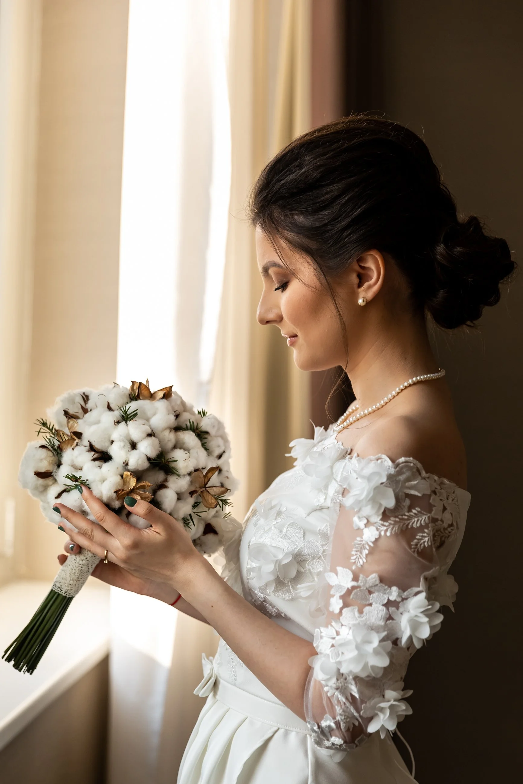 A bride in a white wedding dress with floral lace sleeves, holding a bouquet of cotton flowers accented with greenery, standing near a window with beige curtains, with her eyes closed and a gentle smile.