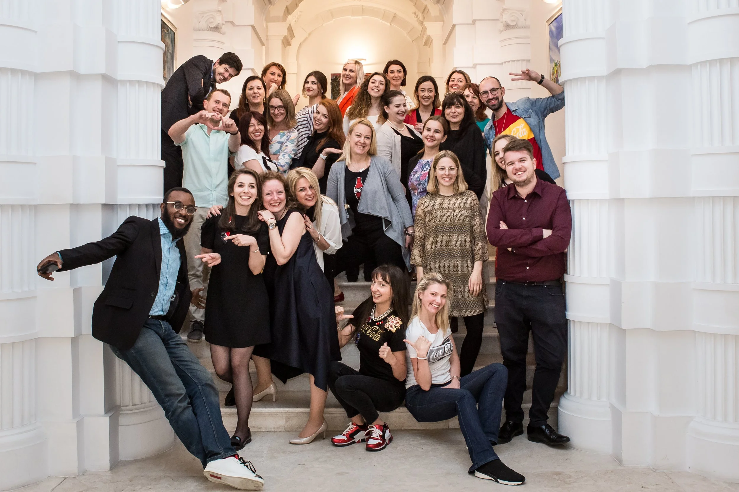 A group of diverse smiling people gathered together on a staircase inside an elegant building with white columns and arches, posing for a photo.