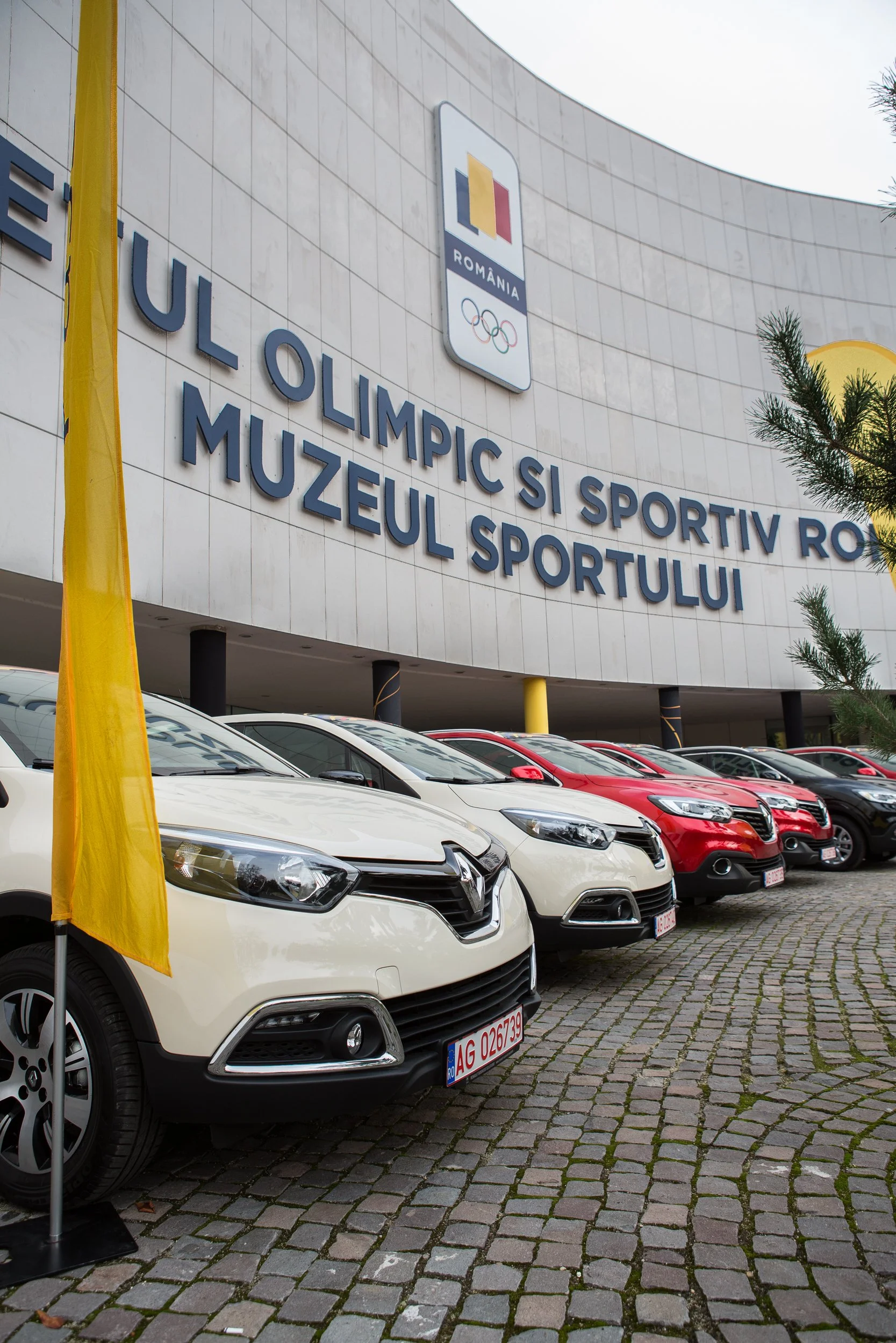 Front view of a sports museum building with cars parked outside. The building has signs indicating it is the Romanian Sports Museum with logos of the Olympic Games. The cars are parked on cobblestone pavement, and there is a yellow flag on the left s