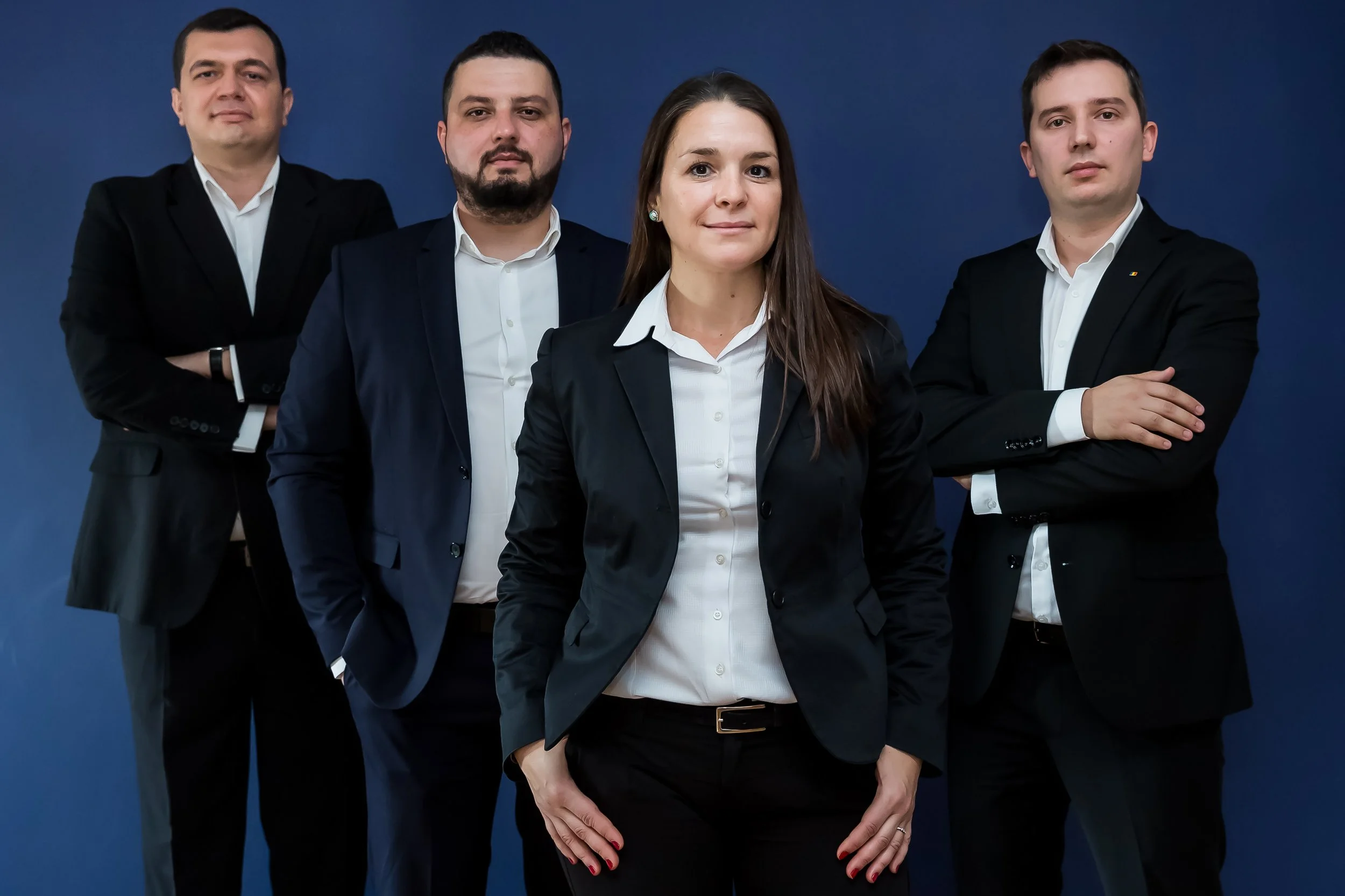 Four professionally dressed individuals, three men and one woman, posing confidently against a dark blue background. The woman stands in the front with one hand in her pocket, while the men stand behind her with arms crossed or relaxed.