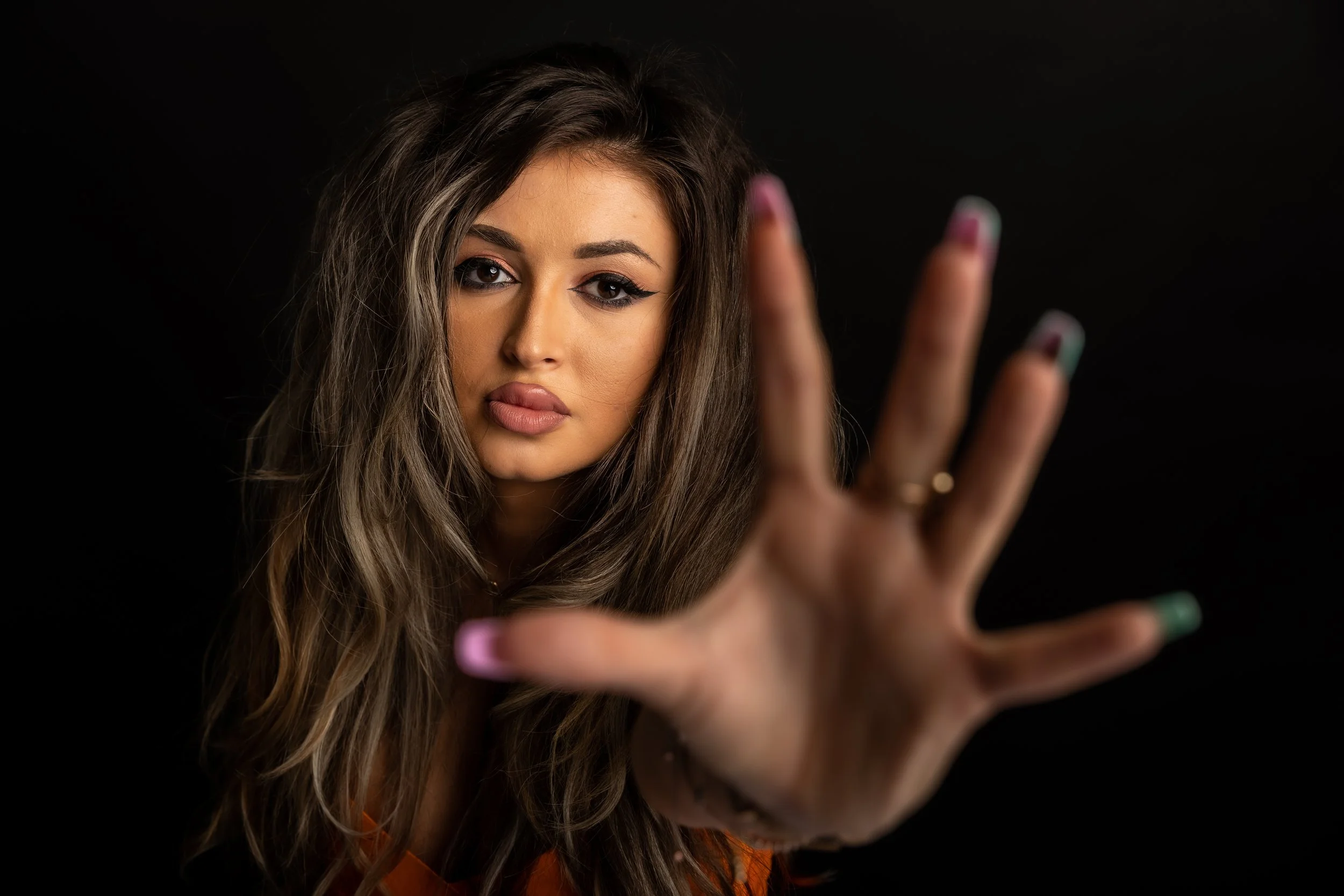 A woman with long wavy hair and makeup taking a selfie with her smartphone, partially extended hand visible, against a black background.
