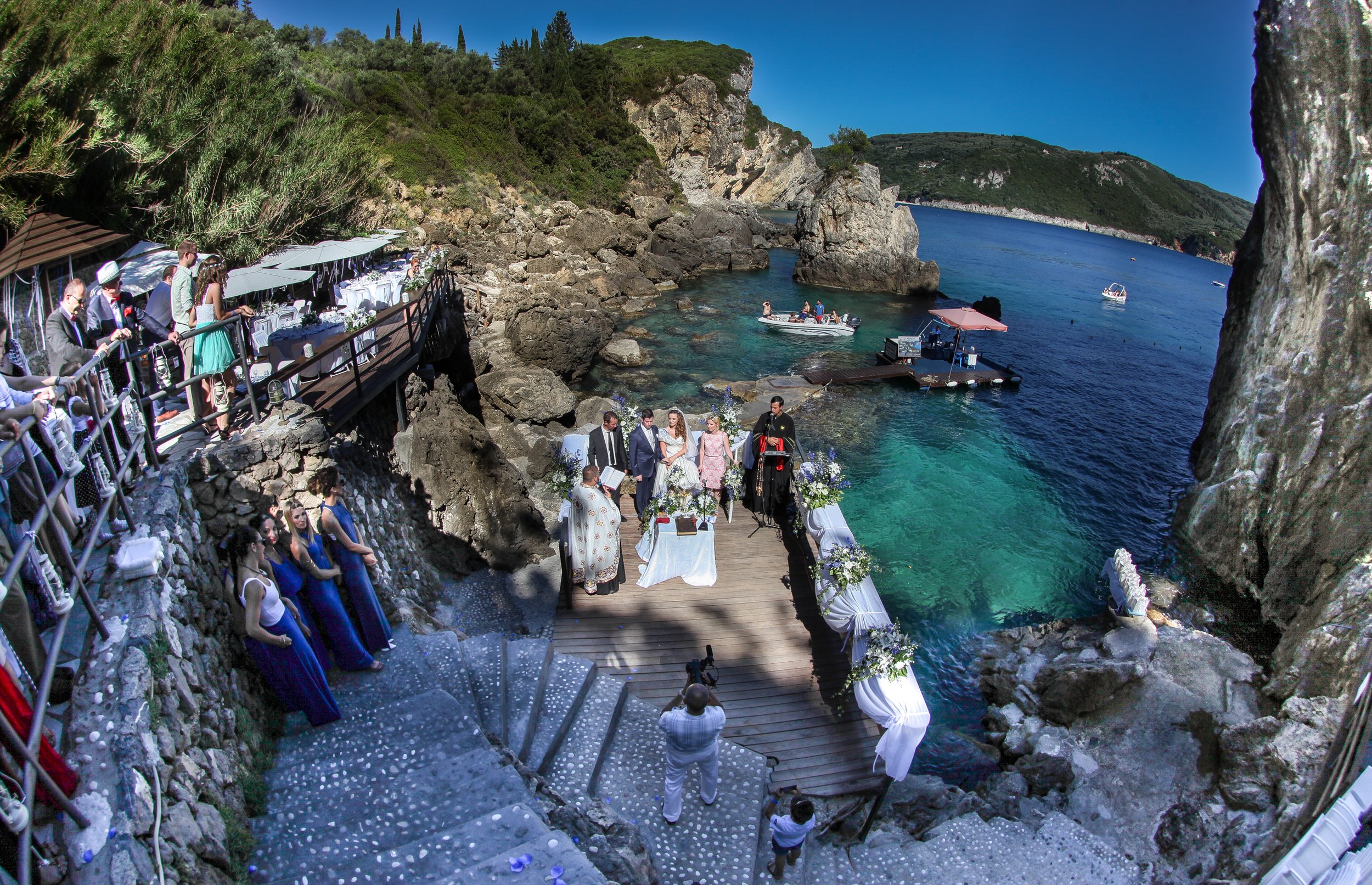 A wedding ceremony on a rocky seaside cliff with guests seated on one side, and an altar set up on a wooden platform overlooking the ocean, with boats in the water and green hills in the background.