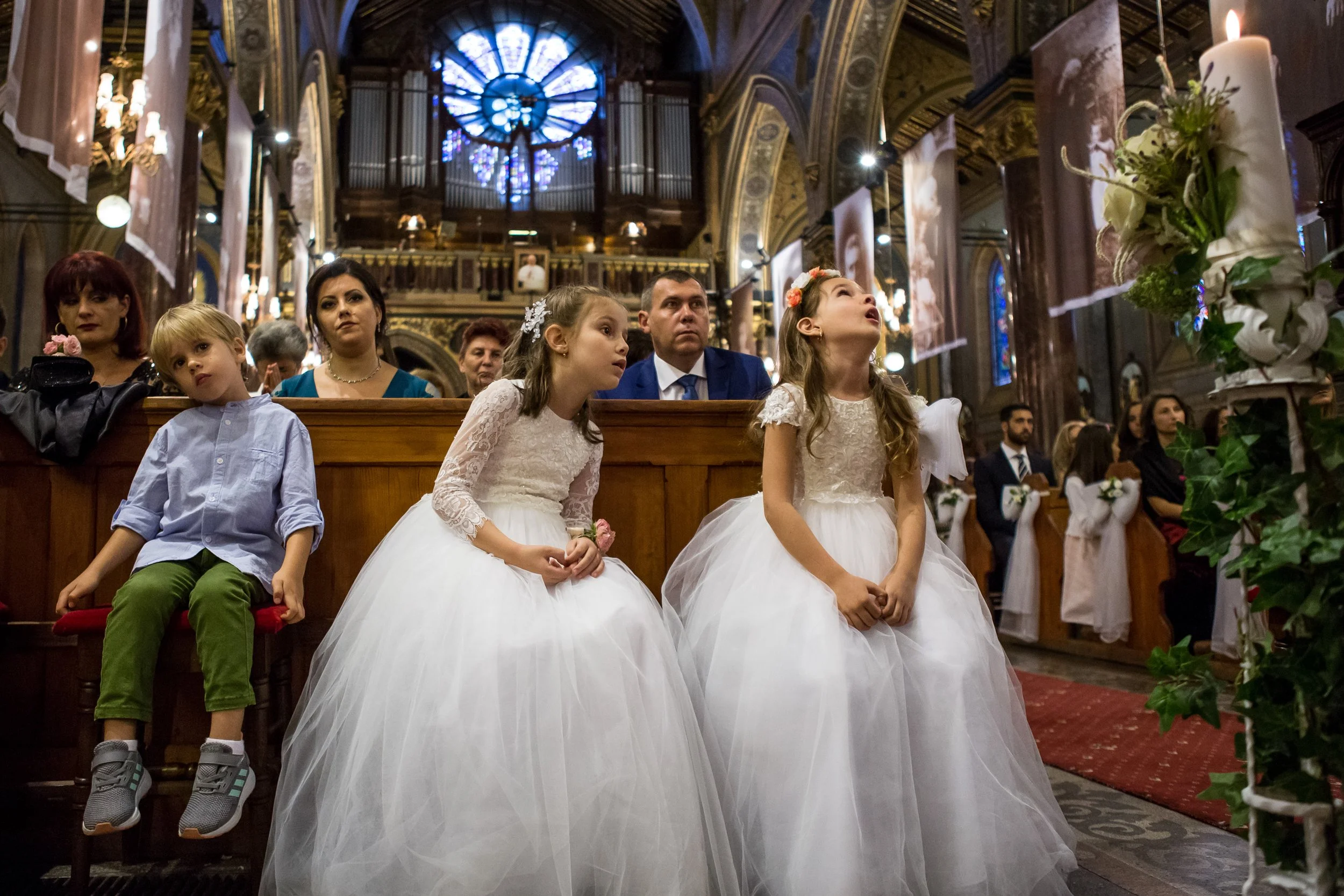 Children dressed as flower girls and young ring bearer sitting in a church during a wedding ceremony, with stained glass windows and ornate architecture in the background.