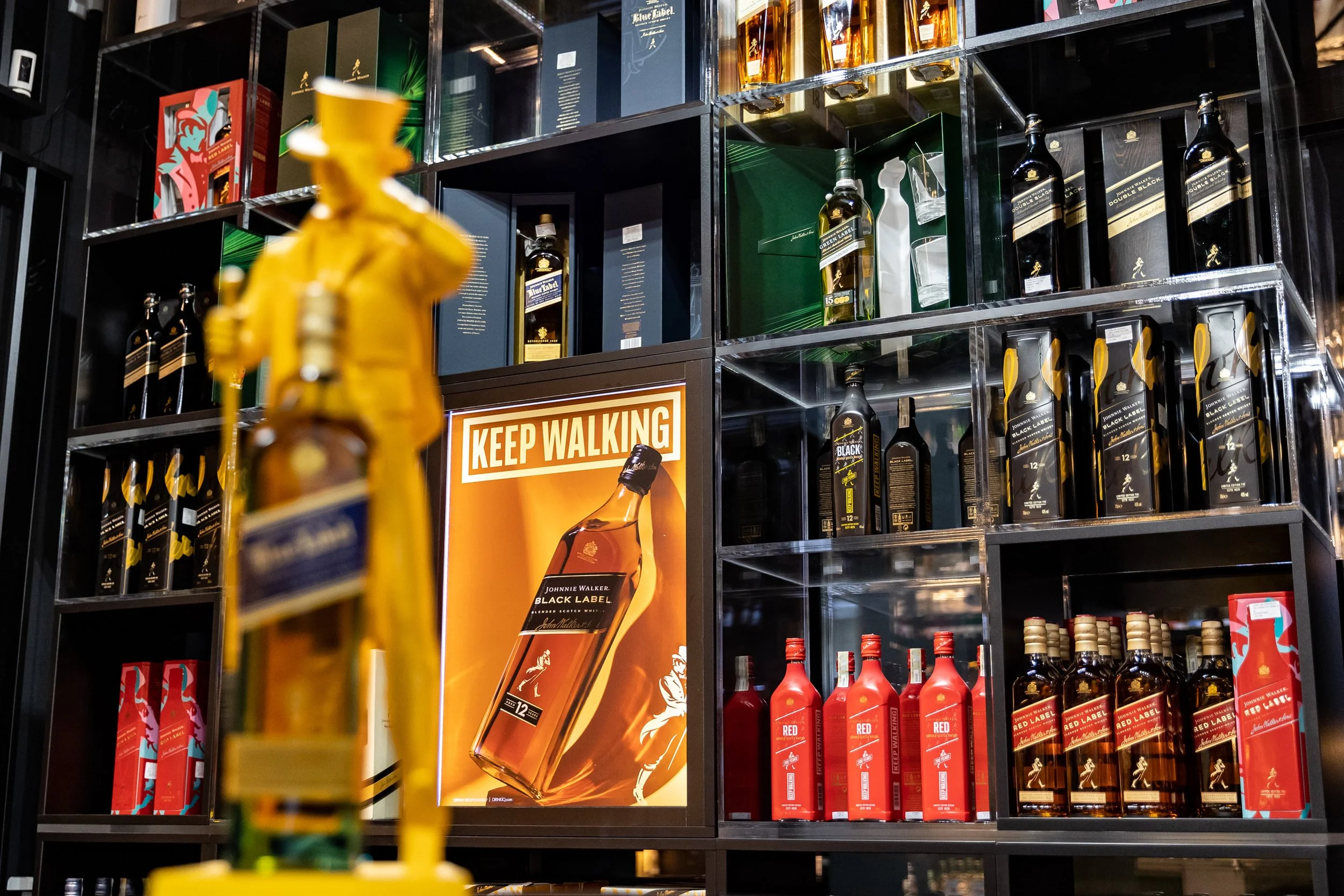 A shelf display of various bottles of whiskey and alcohol in a store, featuring brands like Johnnie Walker Black Label and Red Label, with a "Keep Walking" sign in the foreground.
